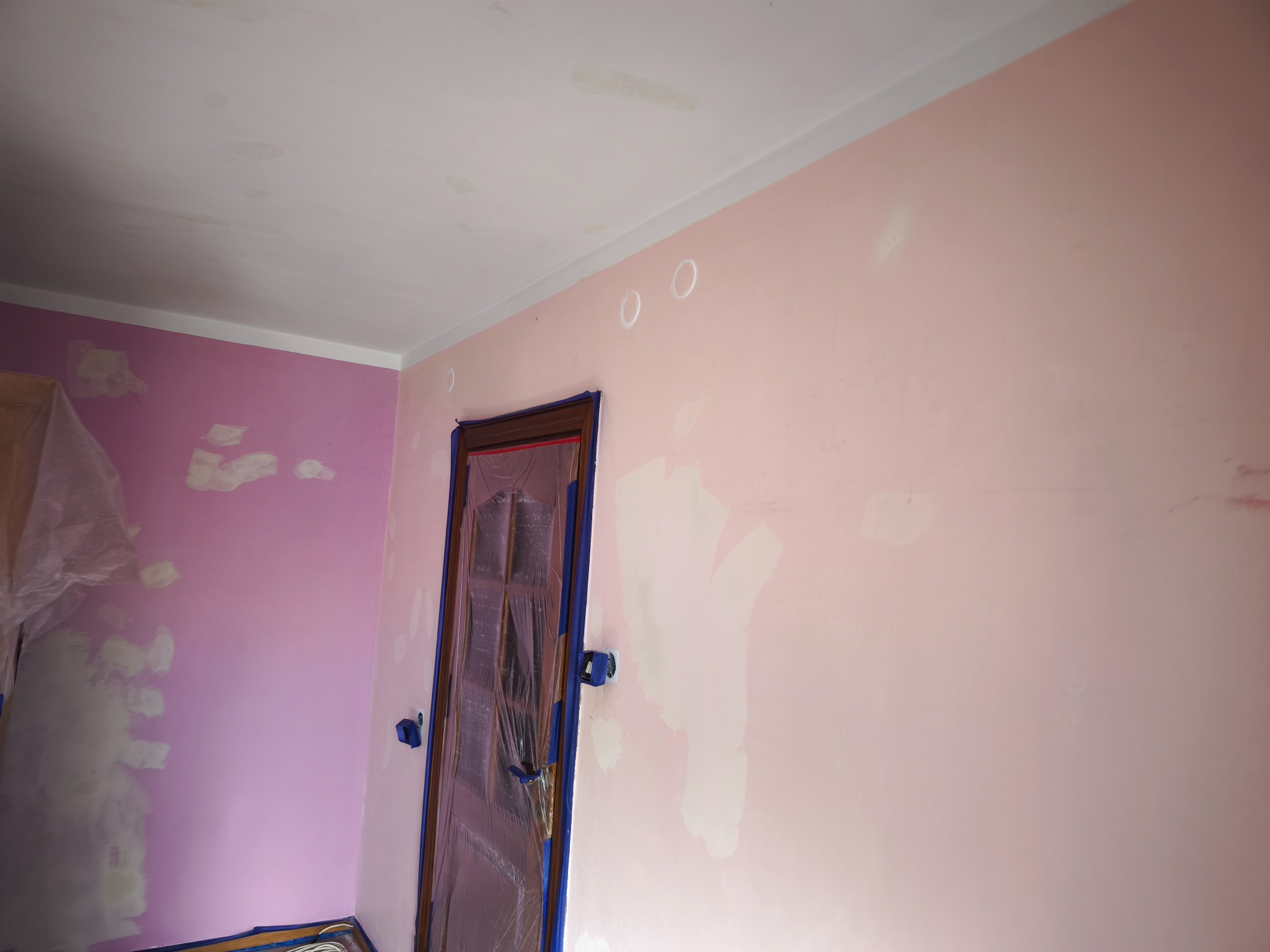 Różowe ściany w trakcie przygotowania do malowania: zagruntowane ubytki, zabezpieczone drzwi folią i taśmą malarską, widoczne ślady po szpachlowaniu na suficie.