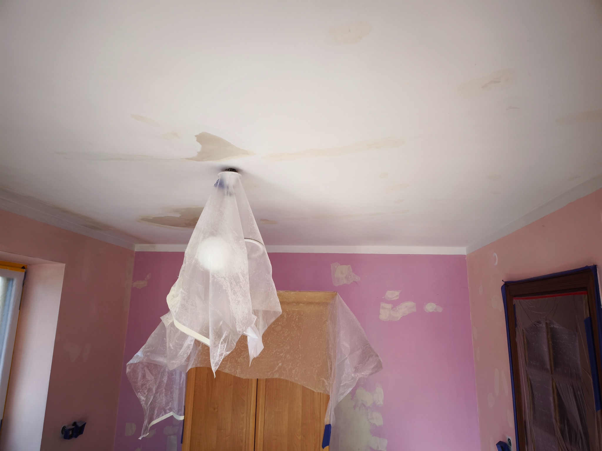 Pokój w trakcie przygotowania do malowania: różowe ściany z widocznymi plamami po szpachlowaniu, zabezpieczona folią lampa sufitowa i okno.