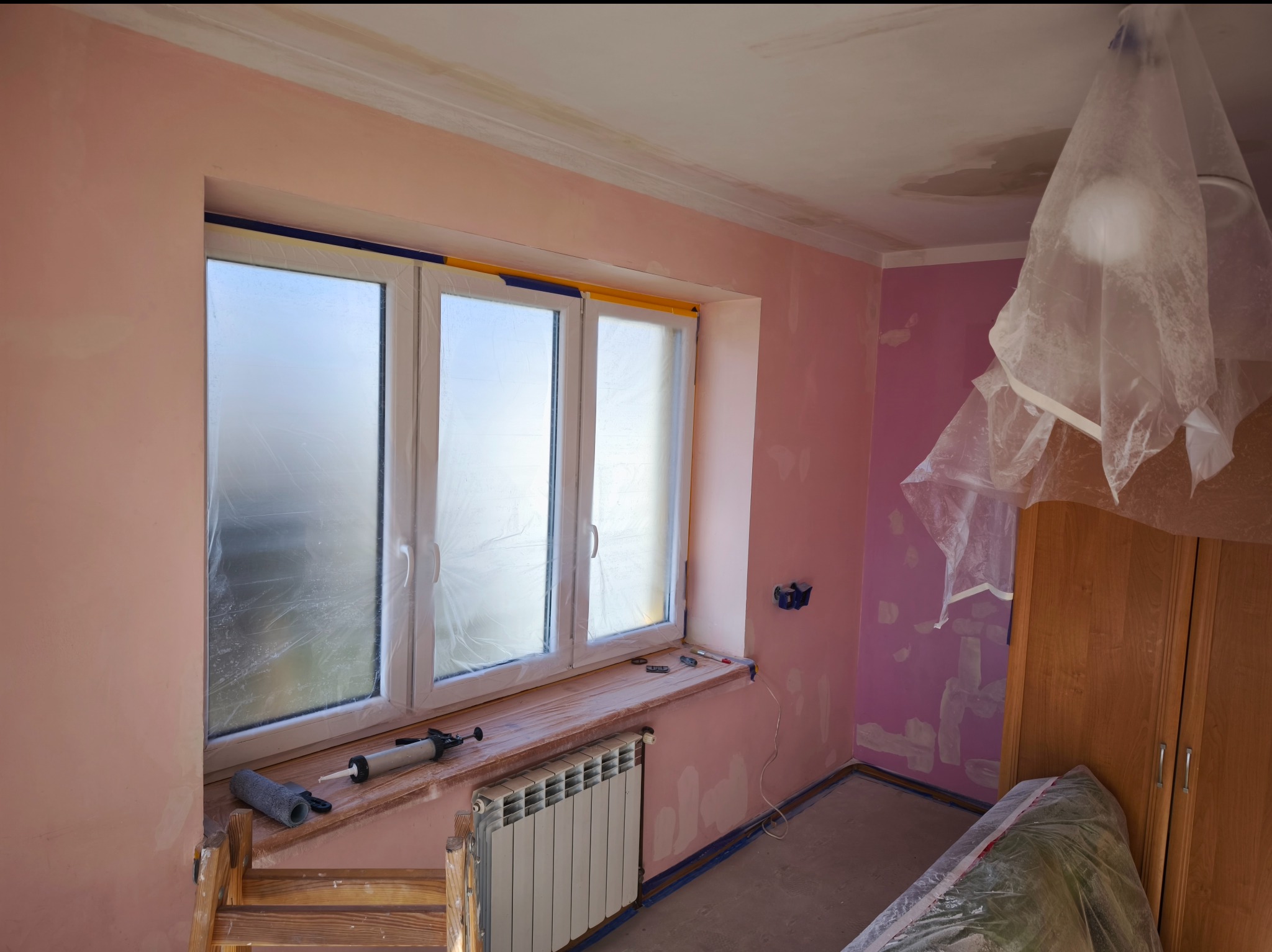 Pokój w trakcie prac wykończeniowych: ściany w odcieniach różu z widocznymi śladami po szpachlowaniu, okno zabezpieczone folią i taśmą, grzejnik pod oknem, drewniana drabina z wałkiem i tubą...