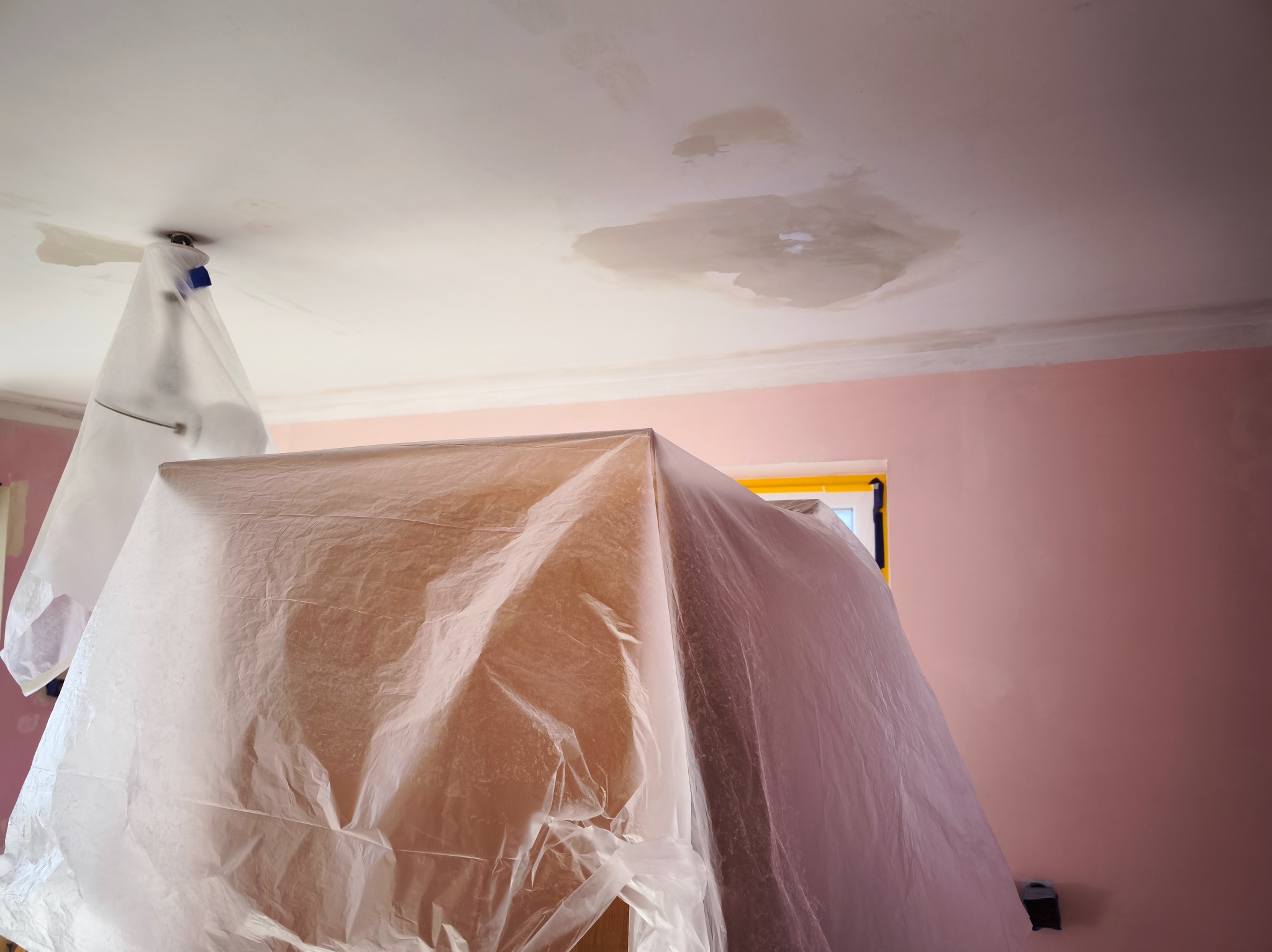 Przygotowanie do prac remontowych: meble zabezpieczone folią malarską, widoczne plamy na suficie i różowe ściany.