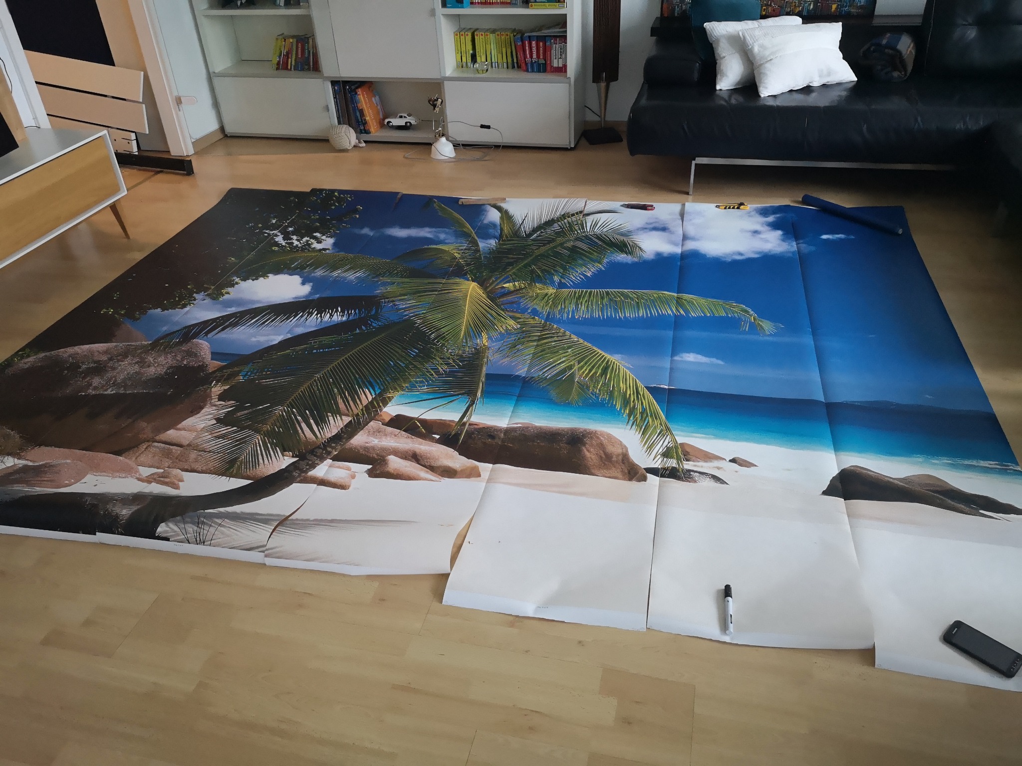 Układanie fototapety z motywem tropikalnej plaży na podłodze, widoczne arkusze do spasowania, marker i telefon komórkowy obok.