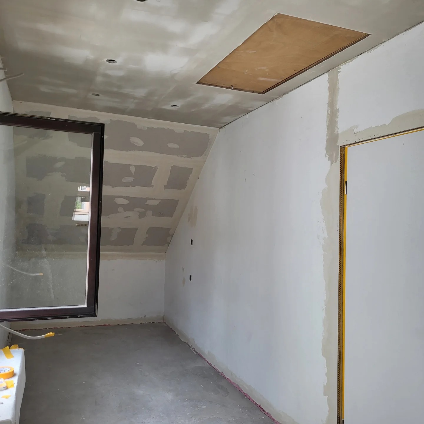 Wnętrze pokoju w Przyszowej w trakcie prac przygotowawczych do wykończenia: ściany po szpachlowaniu, widoczne łączenia płyt gipsowo-kartonowych, surowy beton na podłodze, okno z brązową ramą...