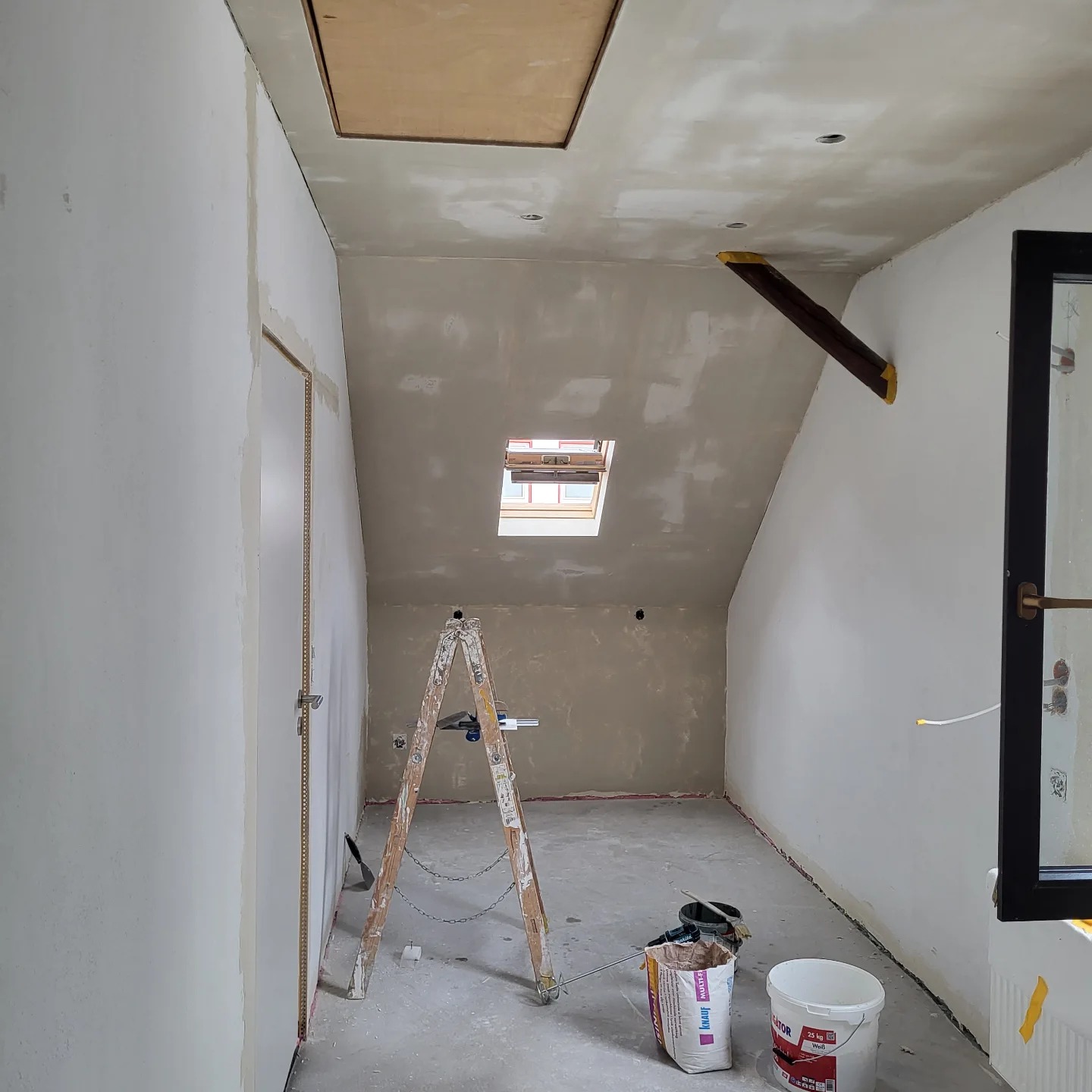 Pokój na poddaszu w trakcie prac wykończeniowych: ściany i sufit po szpachlowaniu, widoczne worki z gipsem, drabina, okno dachowe i fragment okna pionowego.