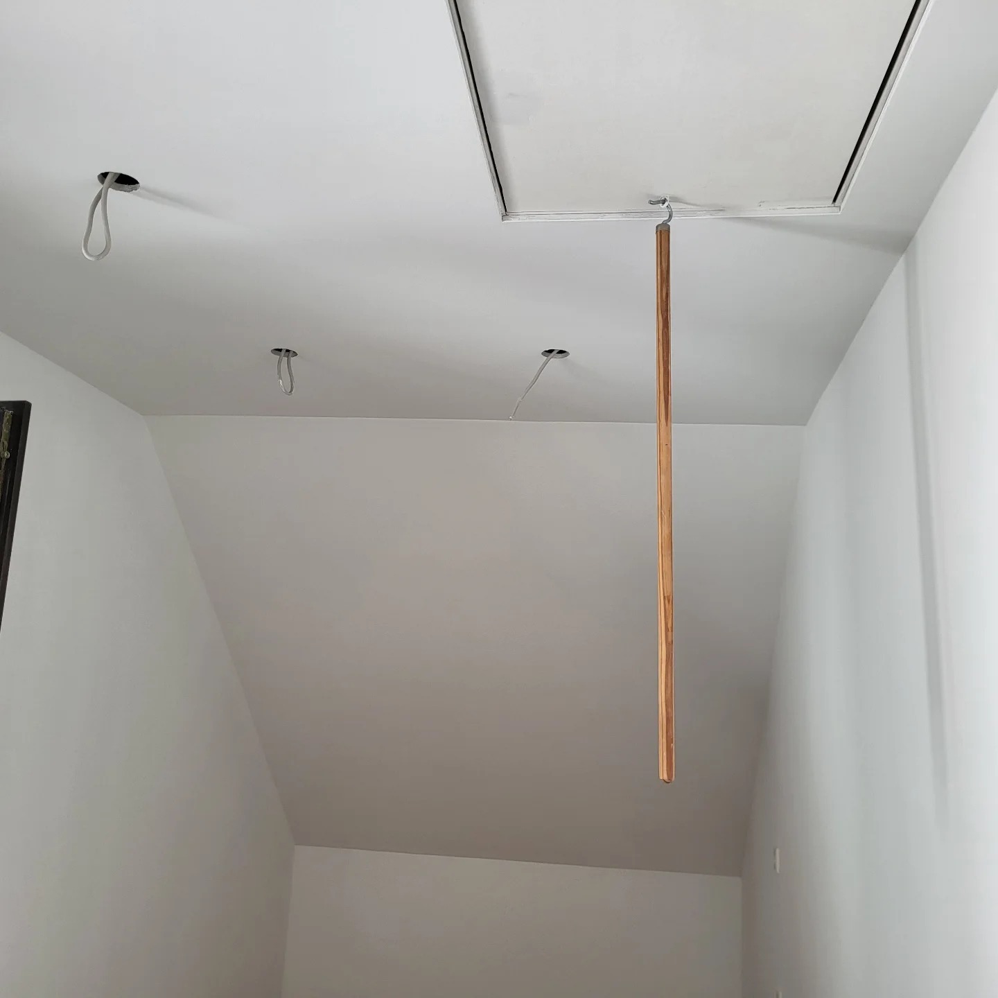 Białe wnętrze poddasza w trakcie malowania, widoczne otwory na oświetlenie, klapa strychowa z haczykiem i wiszącą listwą, surowe ściany.