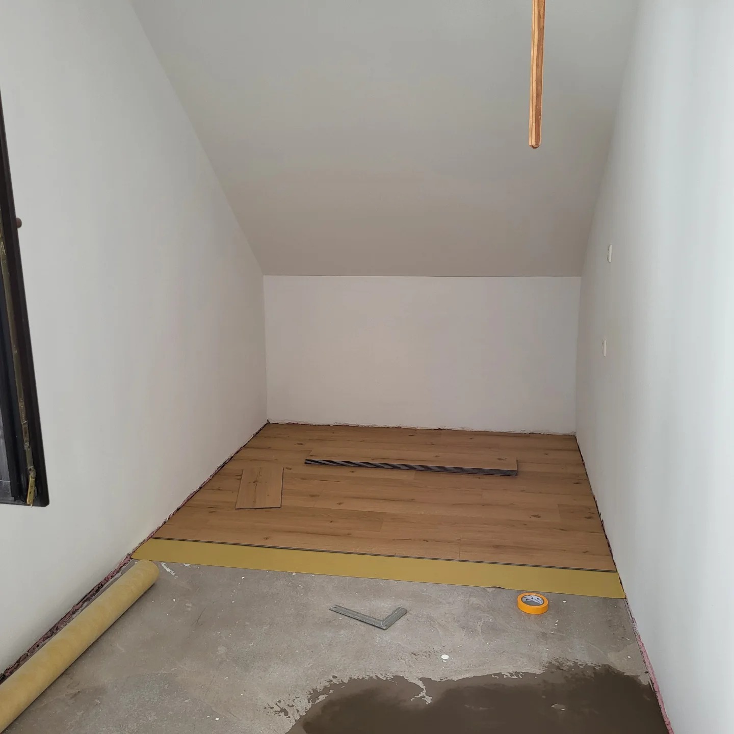 Proces układania paneli podłogowych w pomieszczeniu ze skośnym sufitem, widoczne narzędzia, rolka podkładu i częściowo ułożone panele.