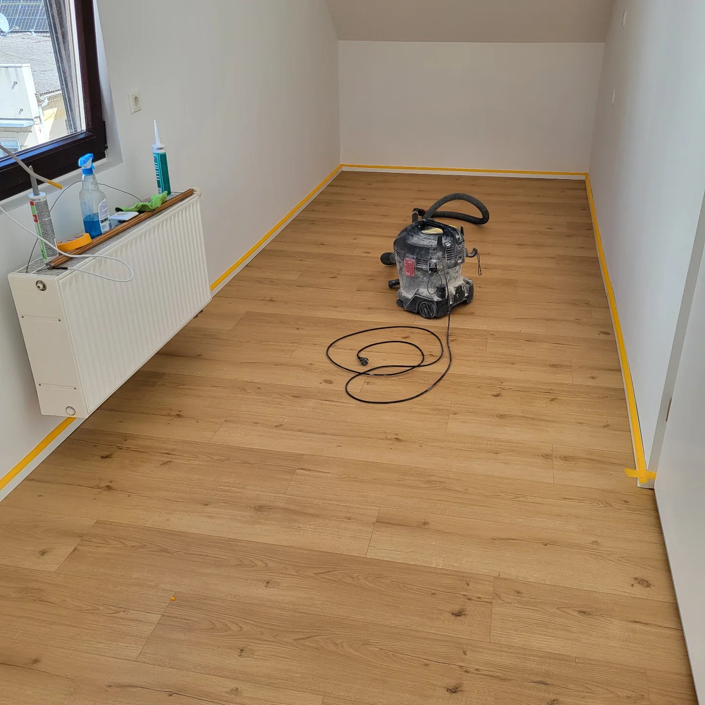 Pokój na poddaszu z nową, jasną podłogą z paneli, zabezpieczony taśmą malarską przy ścianach, z odkurzaczem przemysłowym na środku i grzejnikiem z akcesoriami malarskimi w rogu.