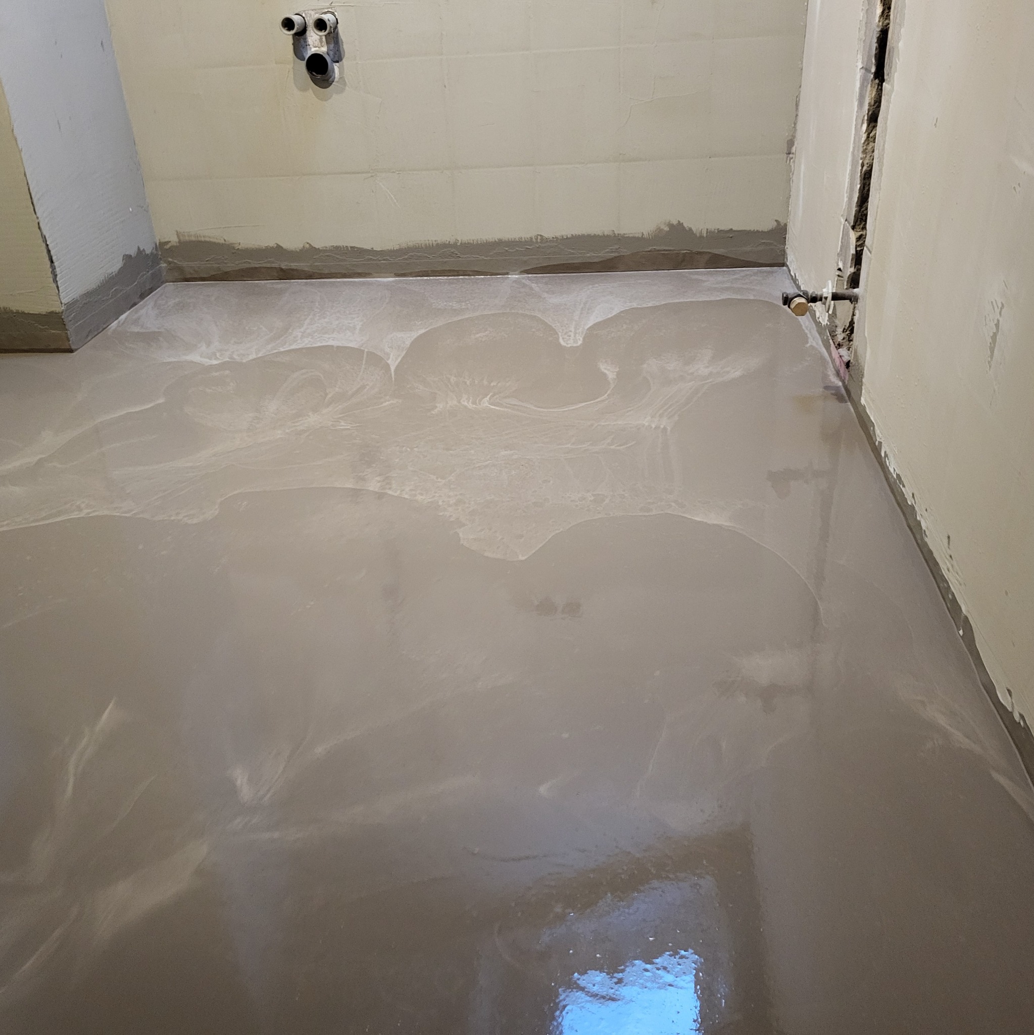 Świeżo wylana, samopoziomująca wylewka podłogowa w łazience, z widocznymi instalacjami hydraulicznymi na ścianie i surowymi ścianami przygotowanymi do dalszych prac wykończeniowych.