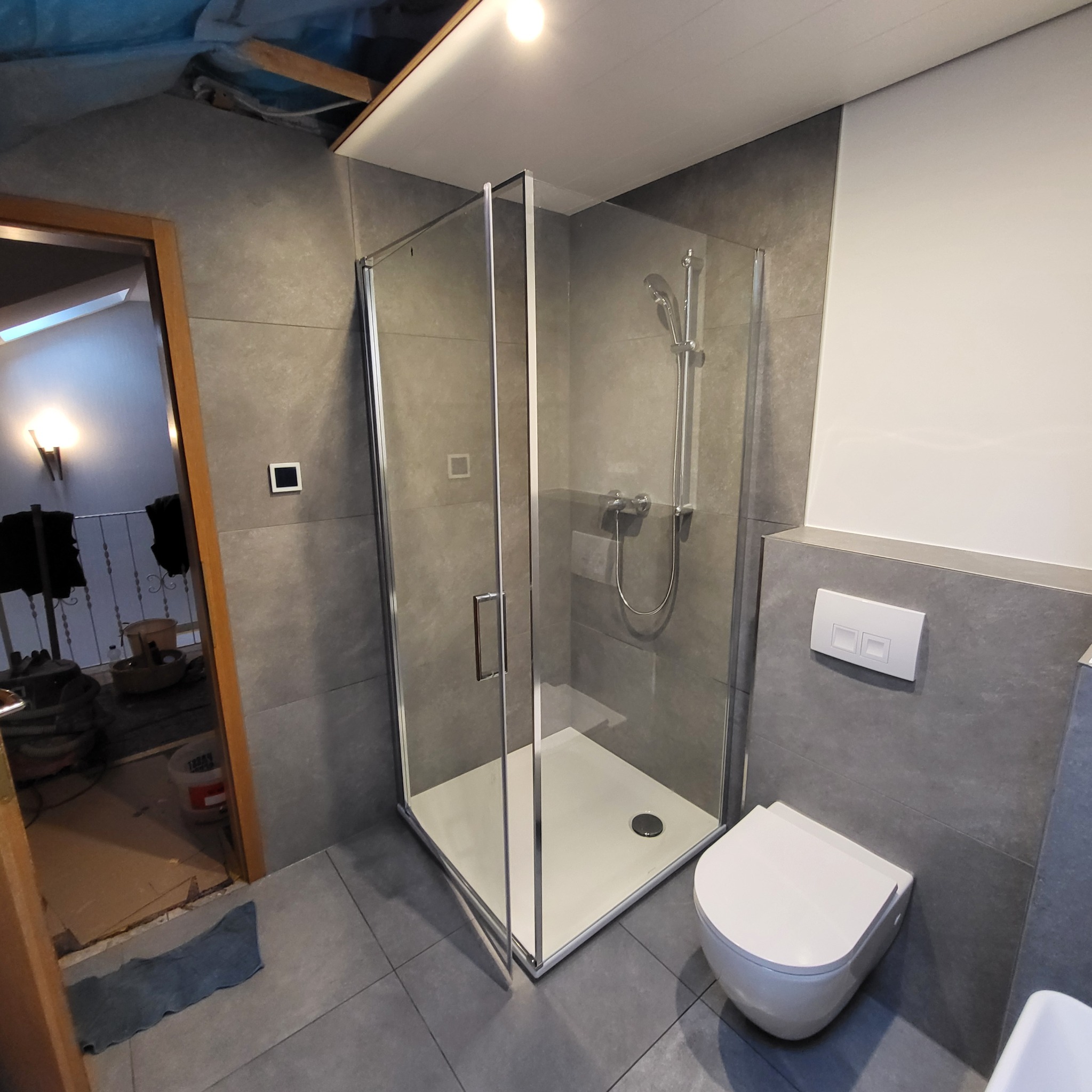 Nowoczesna łazienka z kabiną prysznicową ze szklanymi ściankami, wiszącą toaletą i szarą płytką na ścianach, widoczne ślady prac wykończeniowych przy futrynie drzwi.