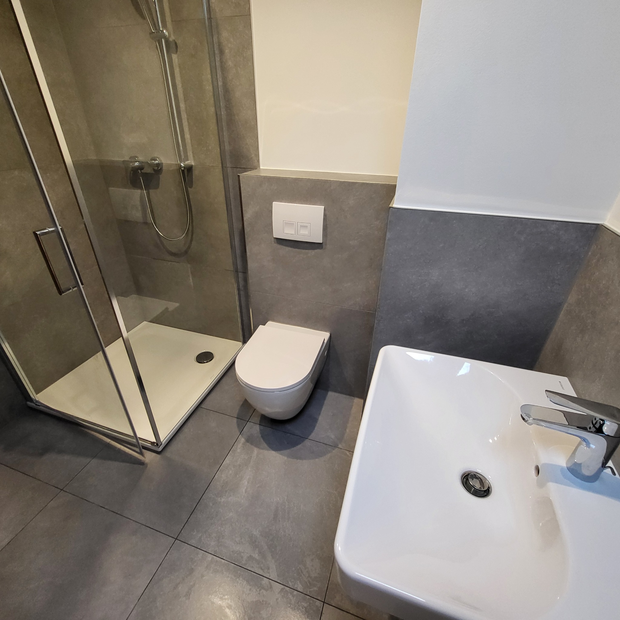 Nowoczesna łazienka z szarą glazurą, kabiną prysznicową z chromowanymi elementami, podwieszaną miską WC i białą umywalką z baterią.