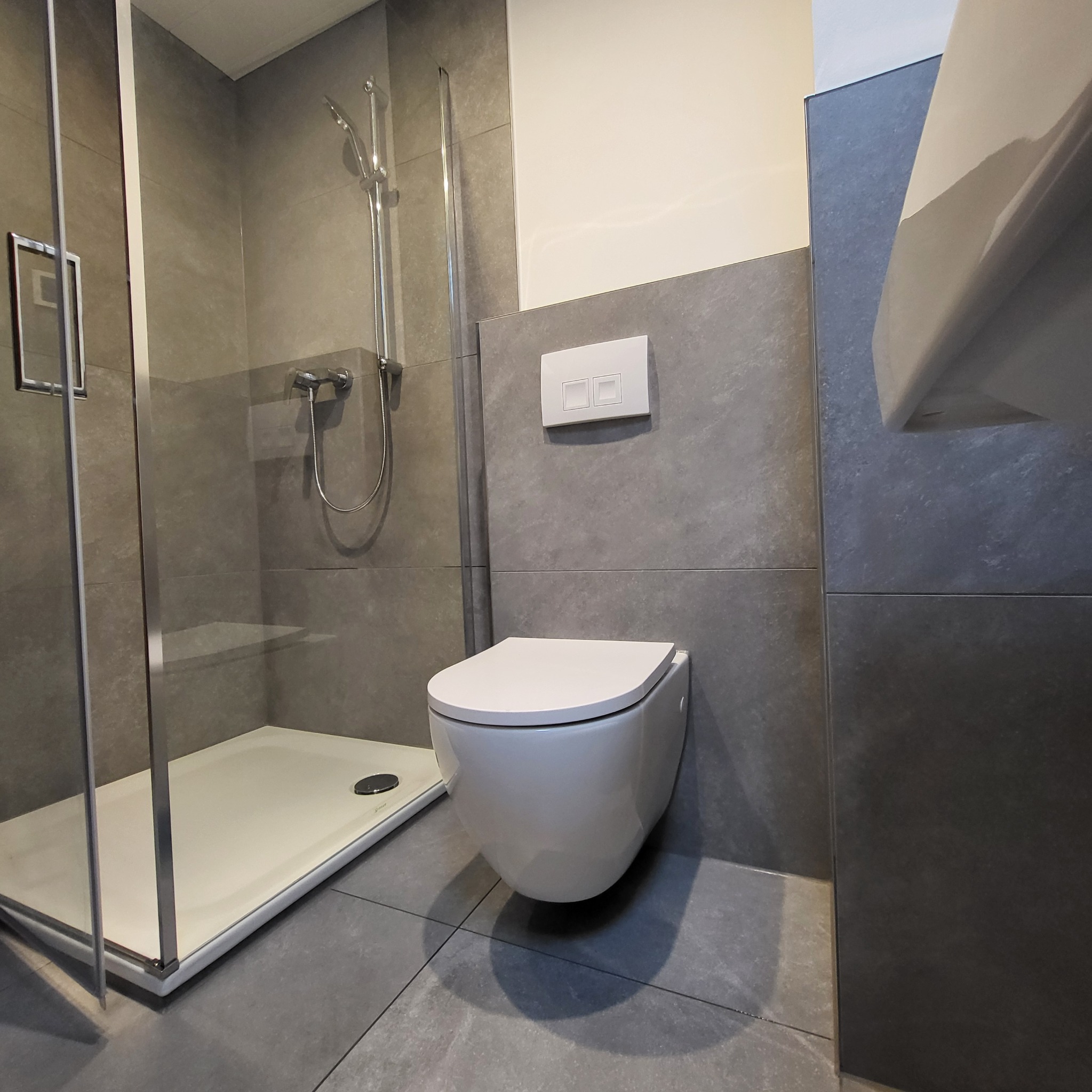 Nowoczesna łazienka z szarą glazurą, kabiną prysznicową typu walk-in i podwieszaną toaletą z białym panelem sterującym na ścianie.