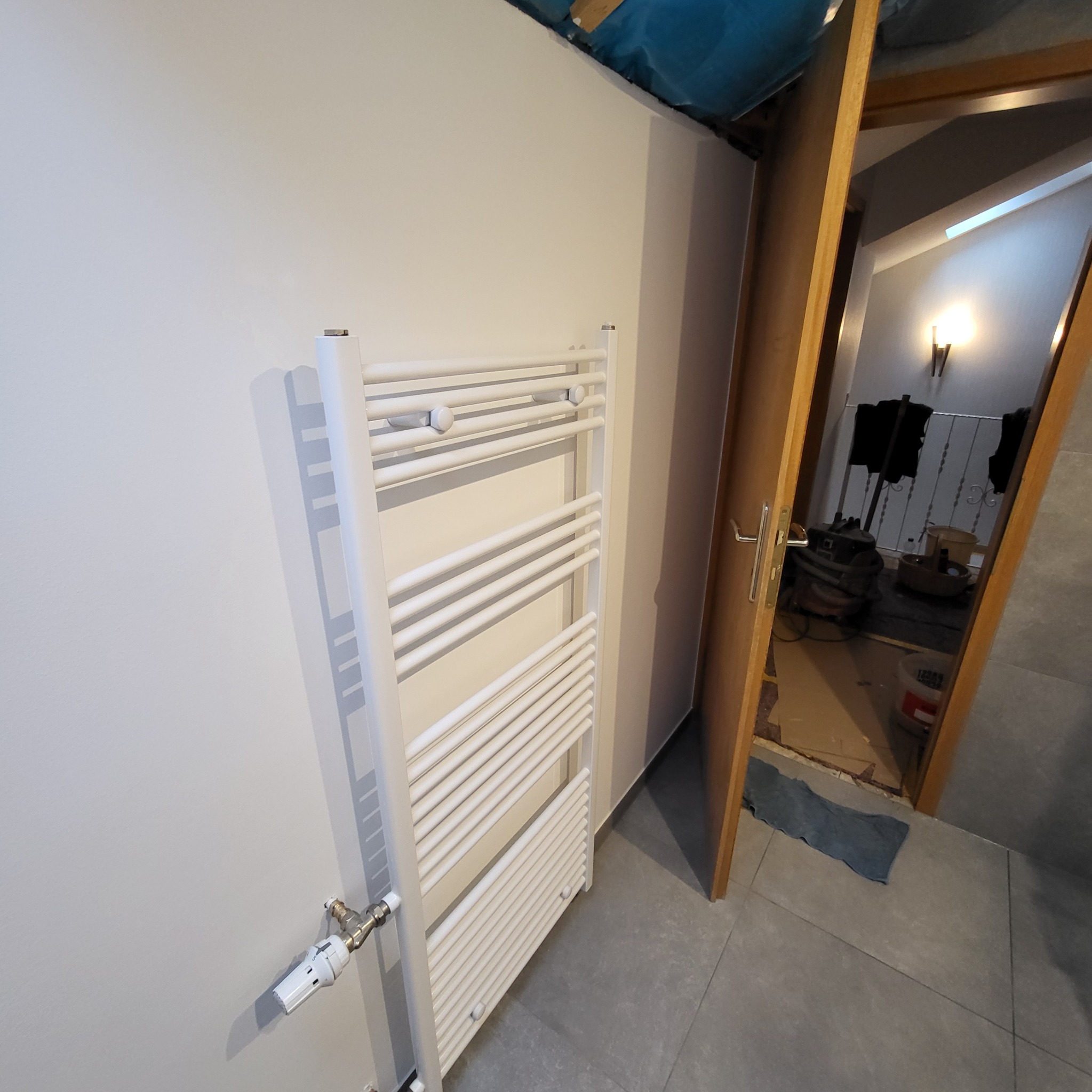 Biały grzejnik łazienkowy drabinkowy zamontowany na ścianie, widoczny zawór termostatyczny, w tle otwarte drzwi i fragment remontowanego pomieszczenia.