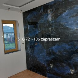 Majster Rob - Łazienka w trakcie wykańczania: ściana z ciemnych, marmurkowych płytek, drewniana podłoga, okno z widokiem na ogród, widoczne instalacje wodne na ścianie, numer telefonu nałożony na zdjęcie.