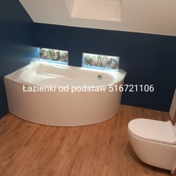 Majster Rob - Nowoczesna łazienka z narożną wanną obudowaną białą zabudową, wisząca toaleta, drewnopodobna podłoga, niebieskie ściany i okno dachowe.