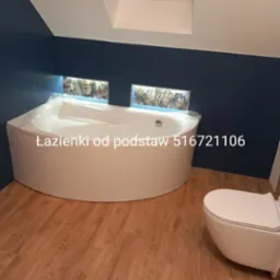 Nowoczesna łazienka z narożną wanną obudowaną białą zabudową, wisząca toaleta, drewnopodobna podłoga, niebieskie ściany i okno dachowe.