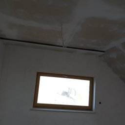 Majster Rob - Wnętrze w trakcie prac wykończeniowych: surowa ściana z oknem w drewnianej ramie, widoczne niedoskonałości tynku na suficie i ścianach, wystające przewody elektryczne.