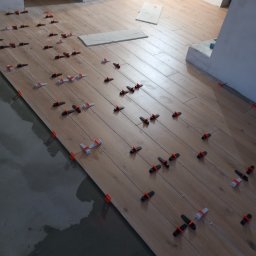 Majster Rob - Układanie drewnopodobnych płytek podłogowych z użyciem klipsów poziomujących, widoczne świeże zaprawy i lampa oświetleniowa w tle.