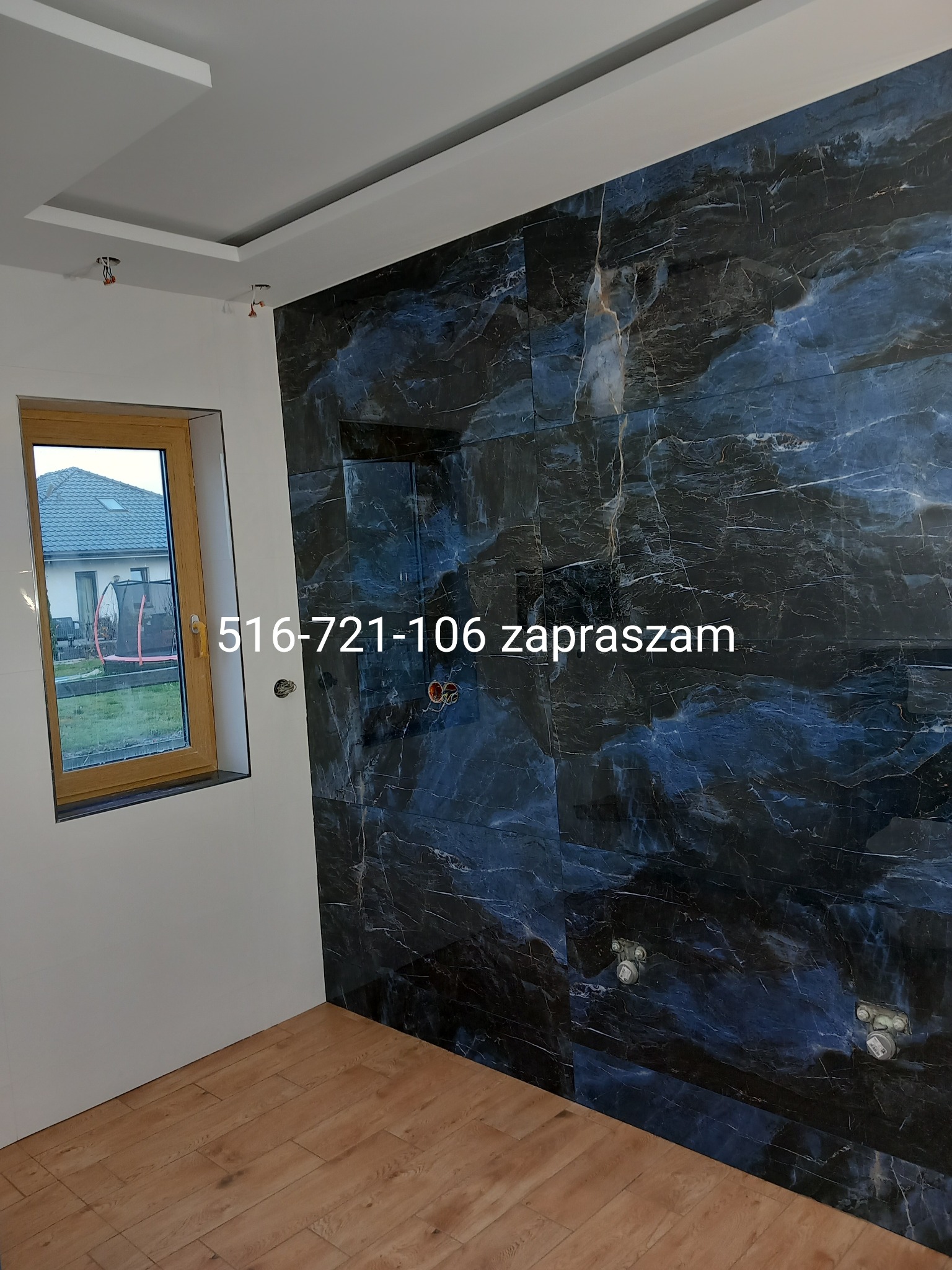 Łazienka w trakcie wykańczania: ściana z ciemnych, marmurkowych płytek, drewniana podłoga, okno z widokiem na ogród, widoczne instalacje wodne na ścianie, numer telefonu nałożony na zdjęcie.
