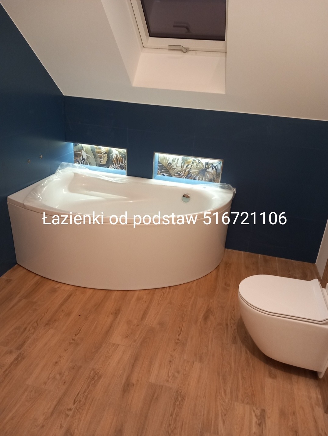 Nowoczesna łazienka z narożną wanną obudowaną białą zabudową, wisząca toaleta, drewnopodobna podłoga, niebieskie ściany i okno dachowe.