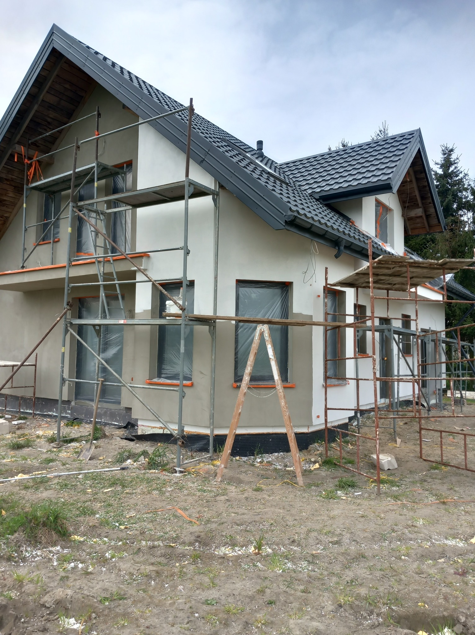 Dom jednorodzinny w trakcie prac elewacyjnych, widoczne rusztowania, częściowo otynkowane ściany, okna zabezpieczone folią.