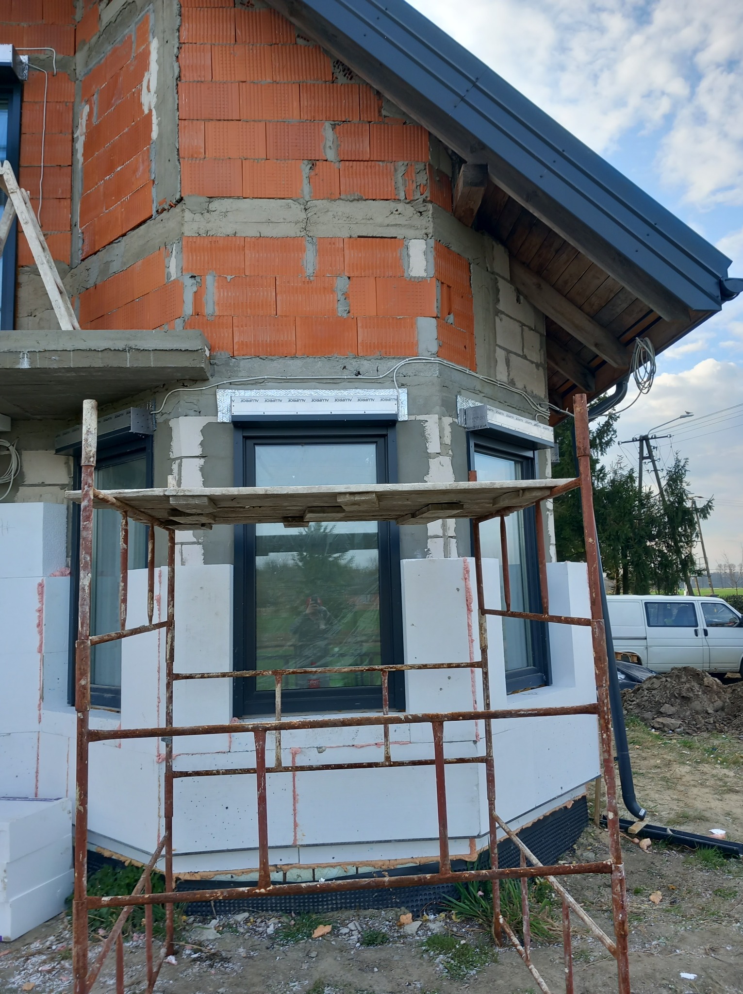 Dom w trakcie budowy: murowana ściana z czerwonej cegły, białe bloczki, zamontowane okna z roletami zewnętrznymi, rusztowanie i widoczny fragment dachu.