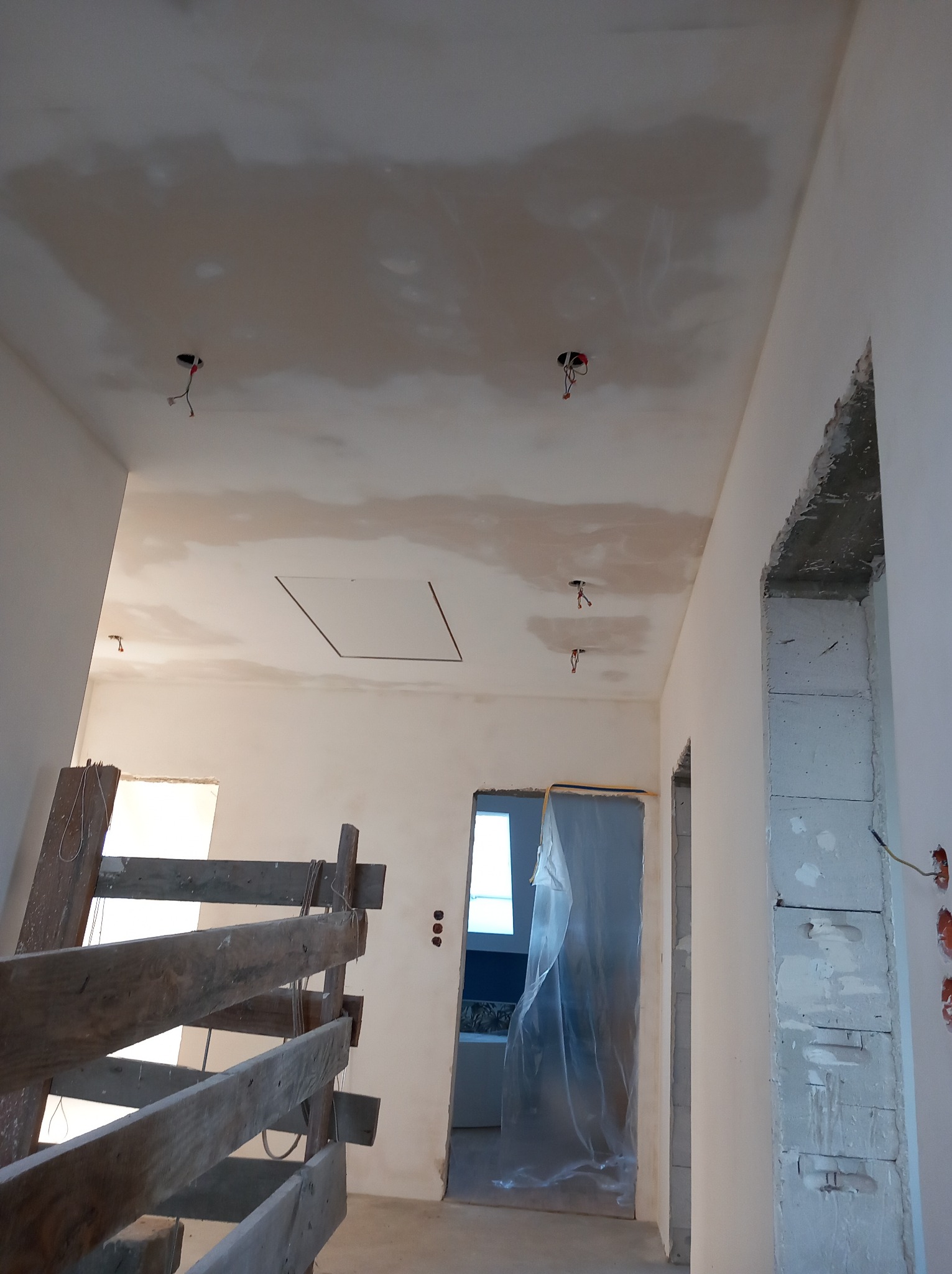 Wnętrze w trakcie tynkowania maszynowego: surowy sufit z widocznymi punktami elektrycznymi, ściany w trakcie wygładzania, drewniane rusztowanie z przodu, widok na sąsiednie pomieszczenie...