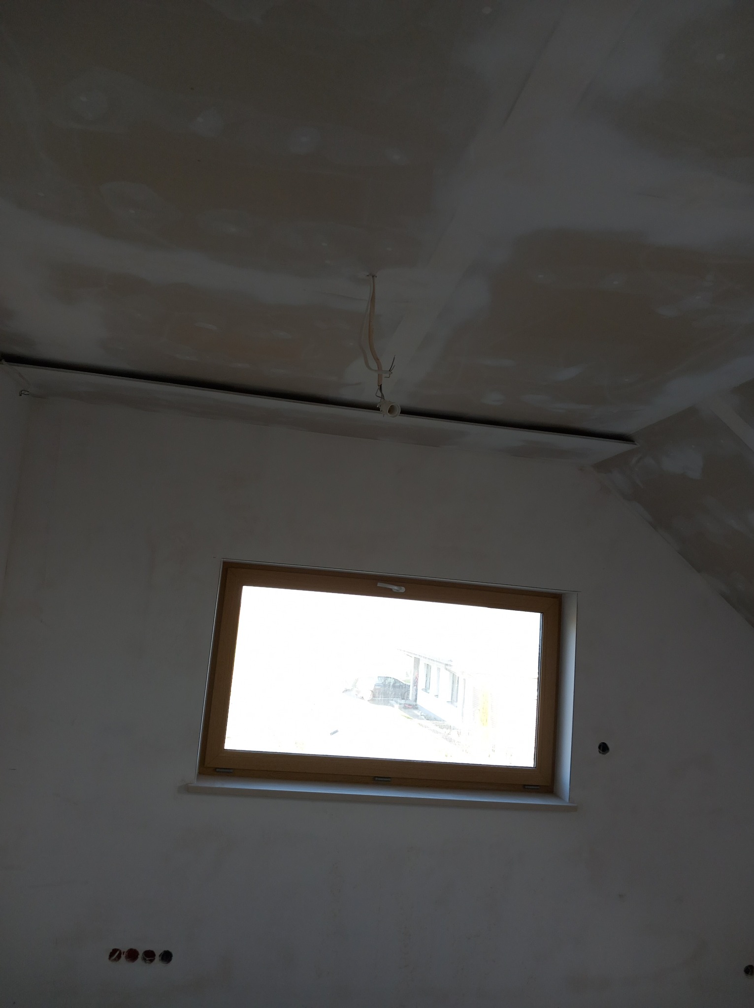 Wnętrze w trakcie prac wykończeniowych: surowa ściana z oknem w drewnianej ramie, widoczne niedoskonałości tynku na suficie i ścianach, wystające przewody elektryczne.