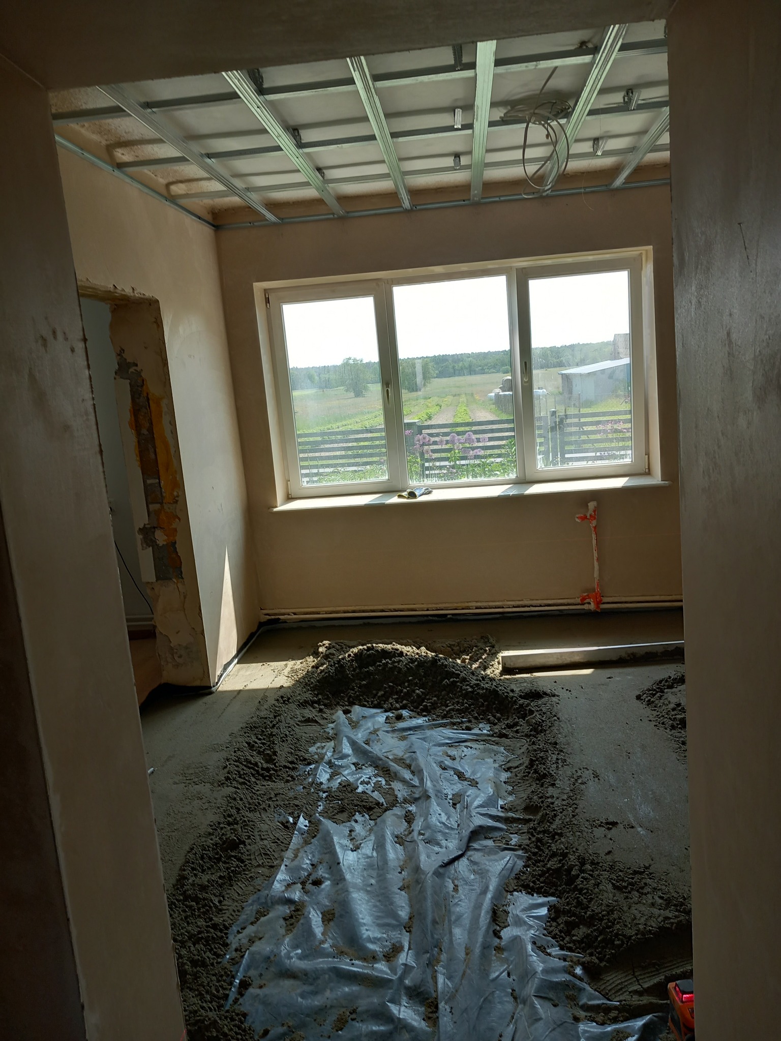 Wnętrze w trakcie prac przygotowawczych do wykończenia: widoczna konstrukcja sufitu podwieszanego, ściany po wstępnym tynkowaniu, podłoga zabezpieczona folią z rozsypanym materiałem budowlanym.