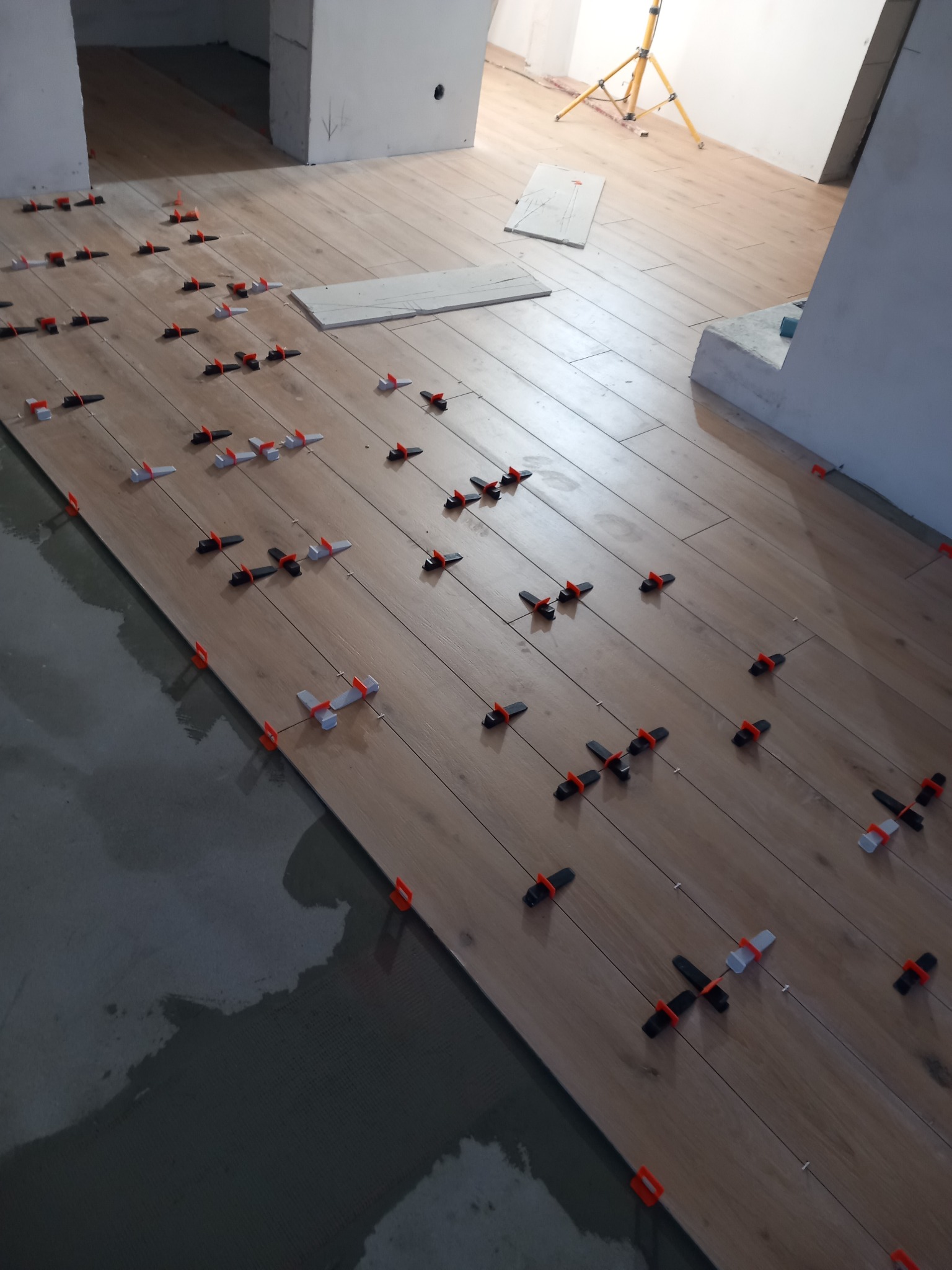 Układanie drewnopodobnych płytek podłogowych z użyciem klipsów poziomujących, widoczne świeże zaprawy i lampa oświetleniowa w tle.