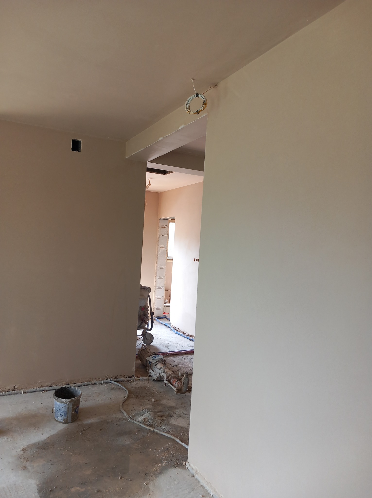 Świeżo nałożony tynk na ścianach w trakcie remontu, widoczna maszyna tynkarska, wiadro i przewody na podłodze, surowy stan wnętrza.