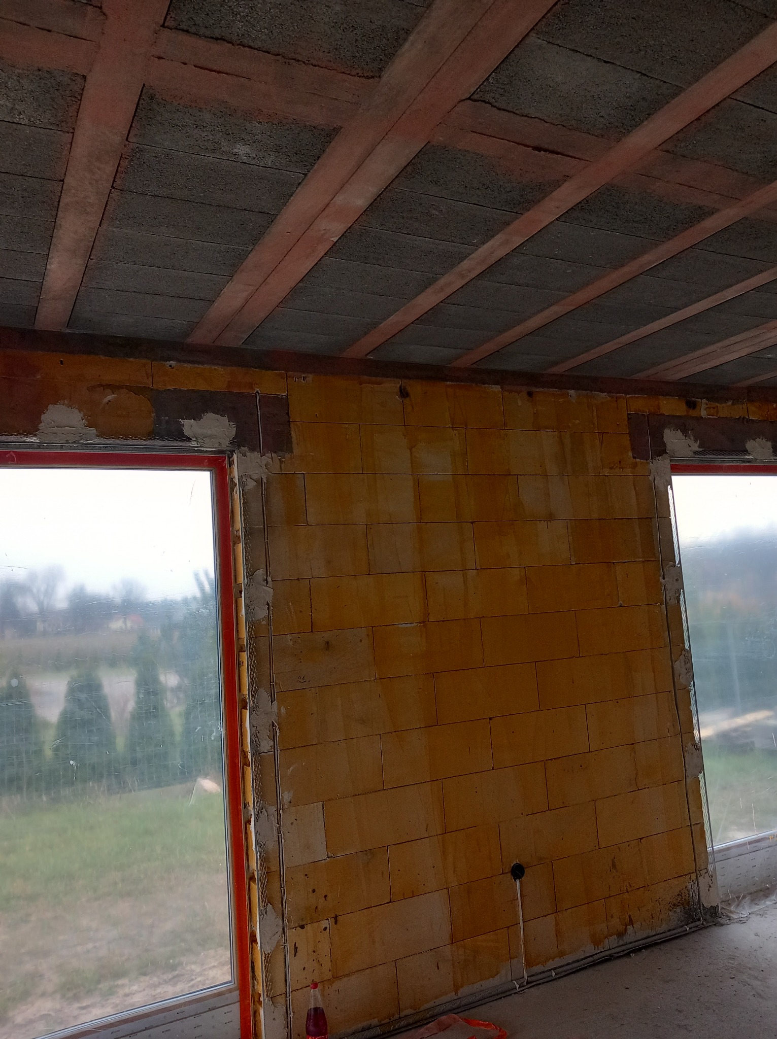 Surowy stan wnętrza: ściana z bloczków, widoczne okno z czerwonym profilem, strop z belek i pustaków, instalacja elektryczna wystająca ze ściany, butelka z napojem na podłodze.