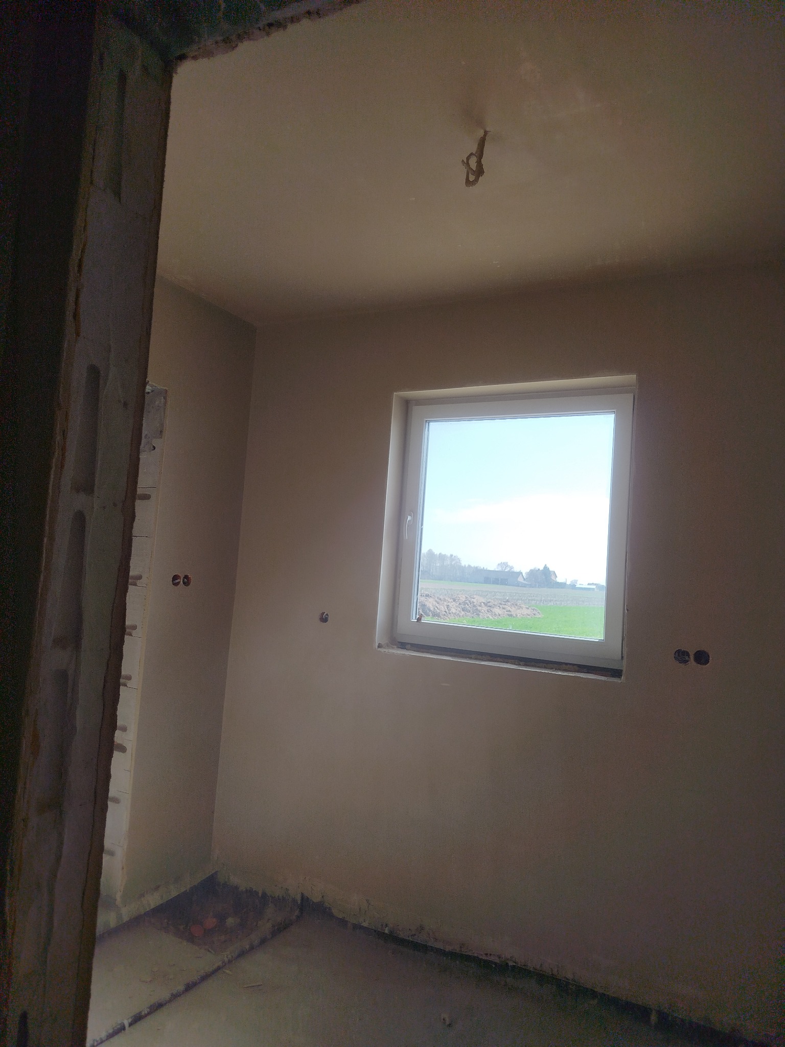 Pokój w trakcie prac wykończeniowych, świeżo otynkowane ściany z widocznymi puszkami elektrycznymi, okno z widokiem na pole, surowa ościeżnica drzwi.