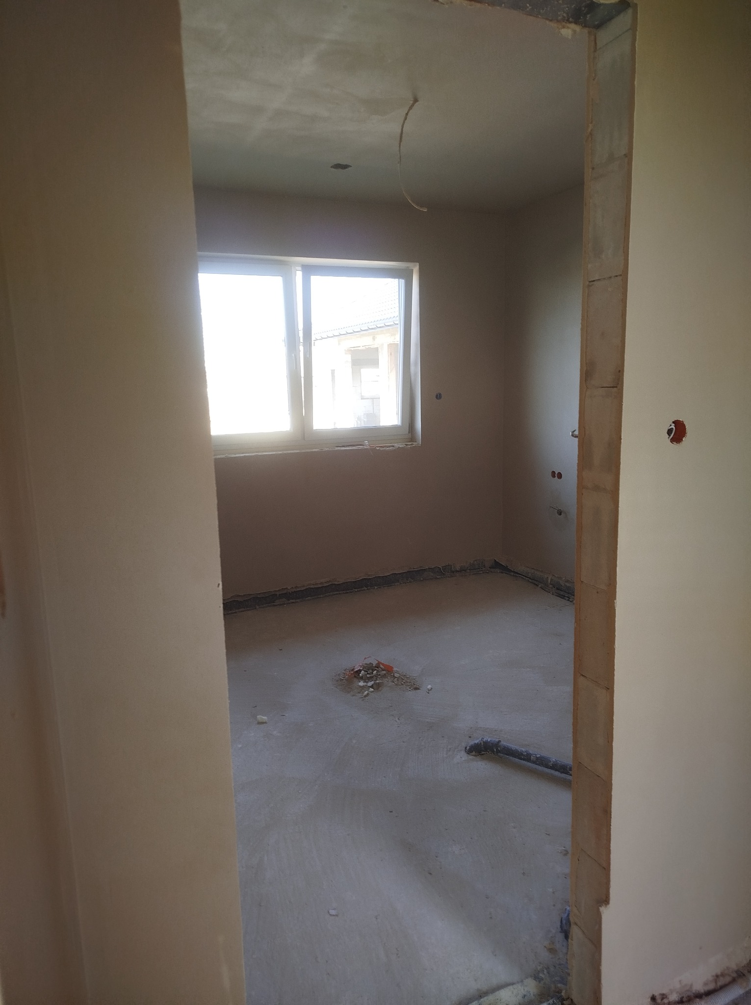 Surowe wnętrze w trakcie wykańczania, widoczne świeżo otynkowane ściany, wylewka na podłodze z wystającymi rurami i instalacją elektryczną, okno z widokiem na budynek.