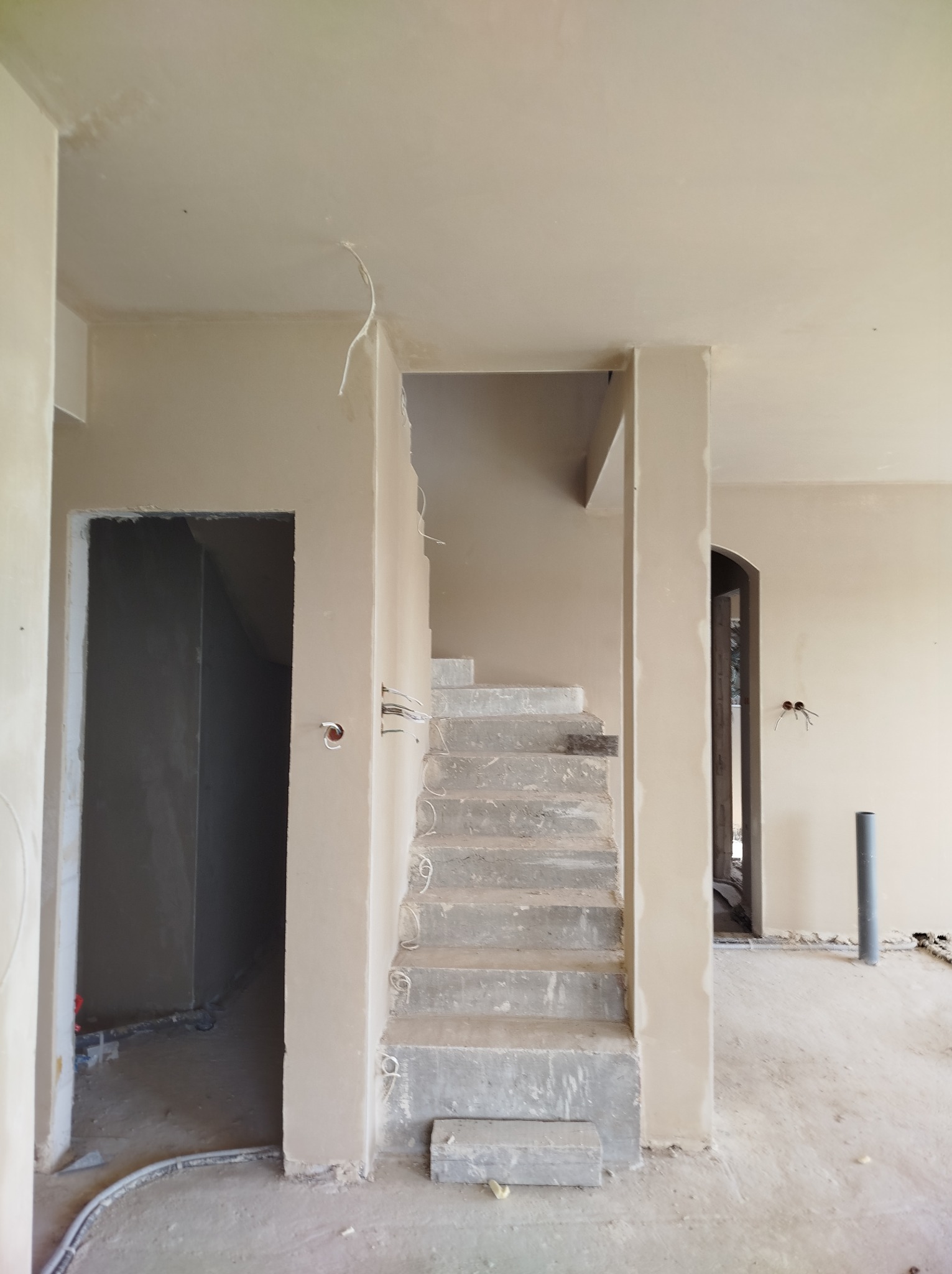 Surowe schody betonowe w trakcie prac wykończeniowych, widoczne przewody elektryczne wystające ze ścian, ściany pokryte świeżym tynkiem, surowy stan wnętrza.
