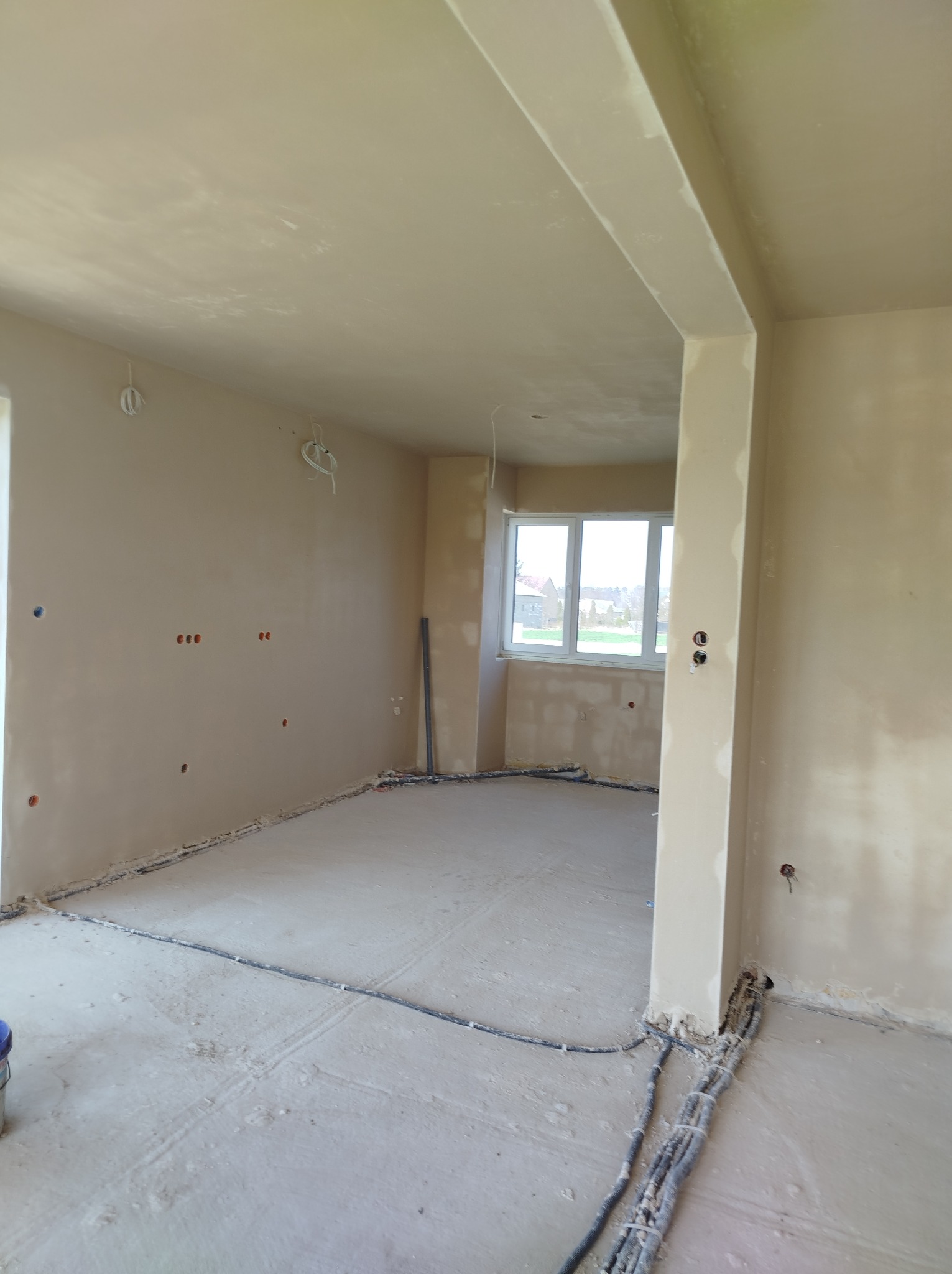 Surowe wnętrze w trakcie prac wykończeniowych: ściany pokryte świeżym tynkiem, widoczne przewody elektryczne wystające ze ścian i podłogi, okno w tle, surowa podłoga z śladami zaprawy.