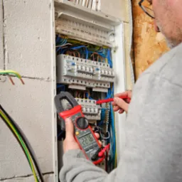 Elektryk mierzy napięcie w rozdzielnicy elektrycznej za pomocą miernika cęgowego, wyświetlającego wartość 227.6, widoczne bezpieczniki Eaton i okablowanie