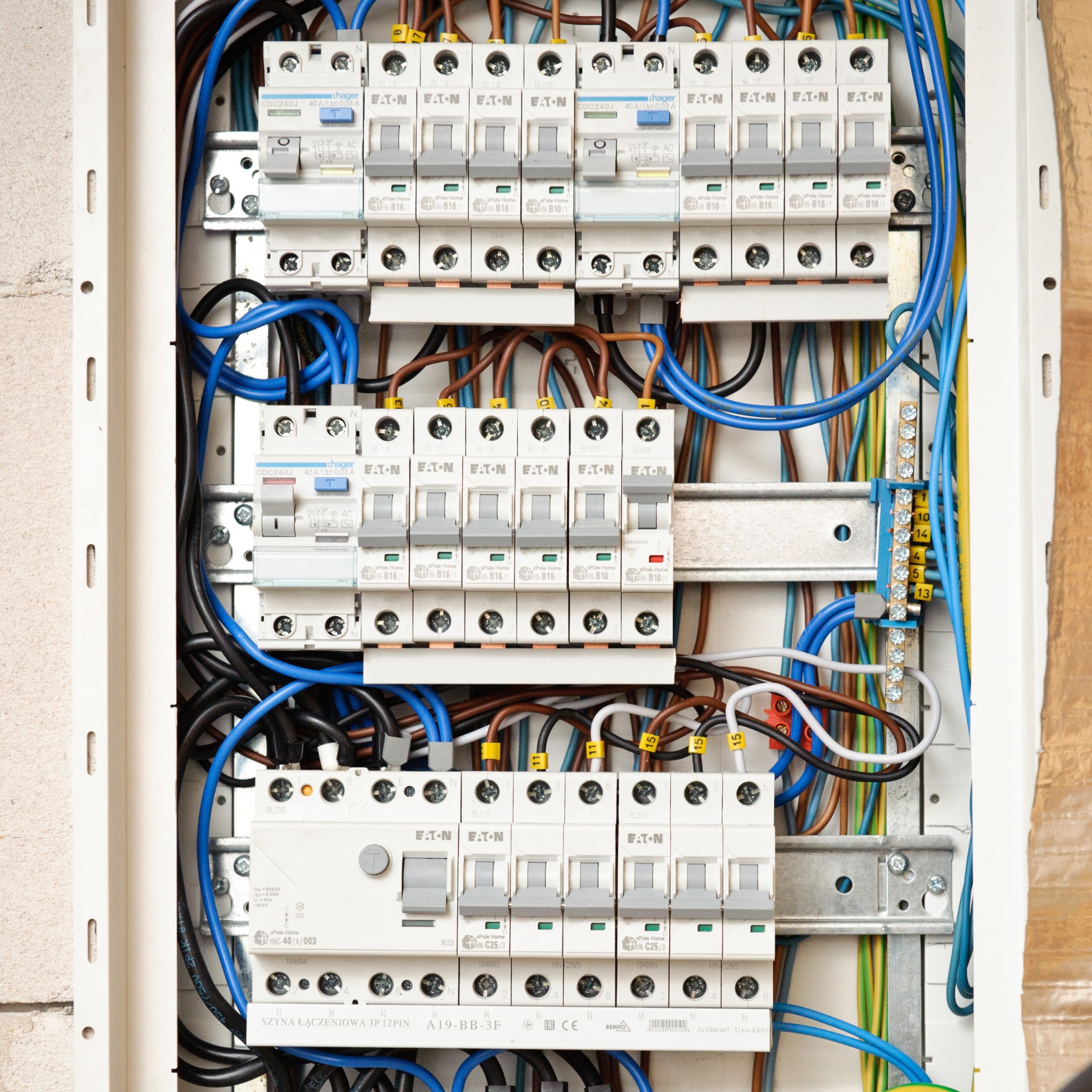 Otwarta skrzynka elektryczna z licznymi bezpiecznikami Eaton i Hager, okablowaniem w kolorach niebieskim, brązowym, czarnym i żółto-zielonym, zamontowana na ścianie z cegły.