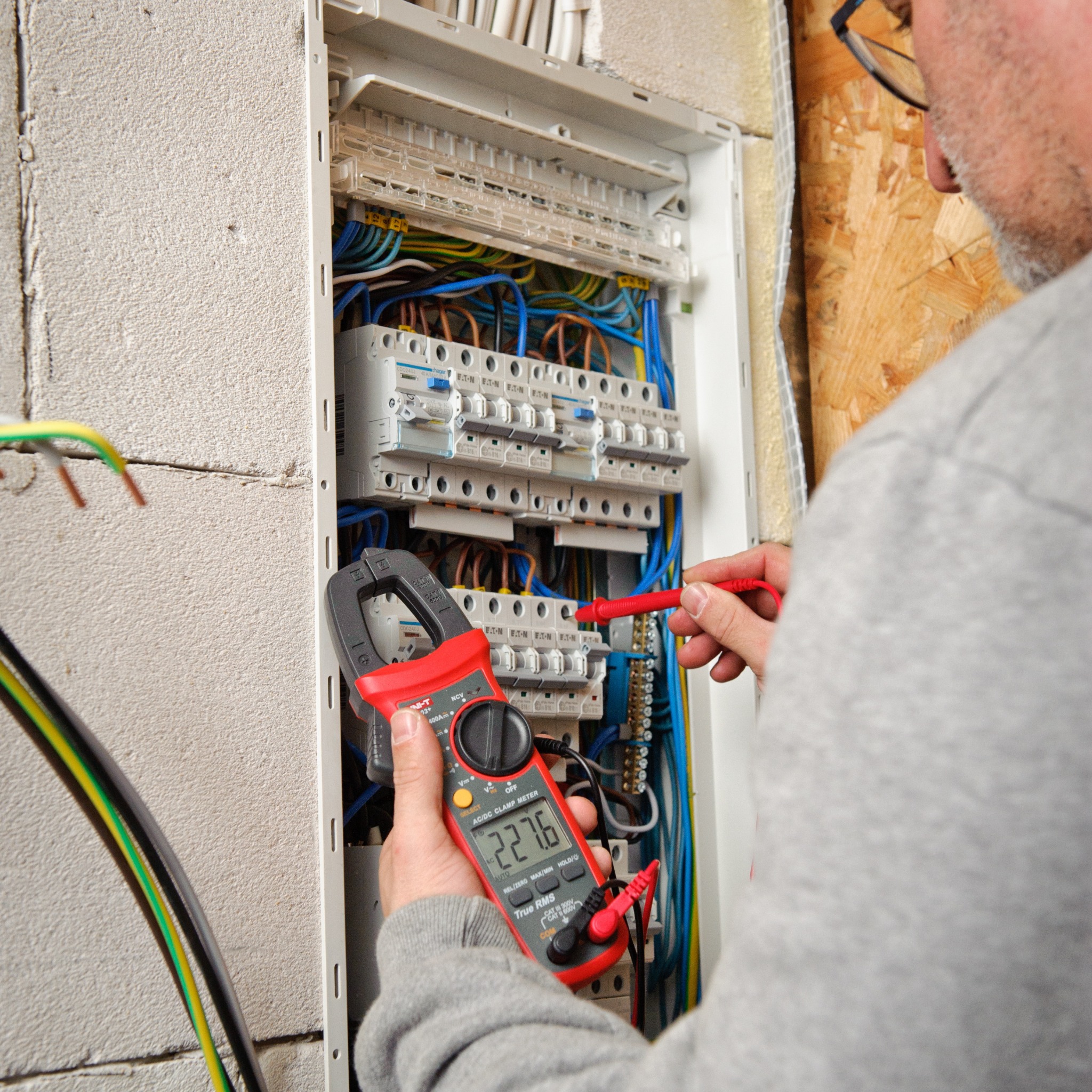 Elektryk mierzy napięcie w rozdzielnicy elektrycznej za pomocą miernika cęgowego, wyświetlającego wartość 227.6, widoczne bezpieczniki Eaton i okablowanie