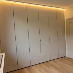 Verdini Andrzej Piotr - Beżowa szafa wnękowa z oświetleniem LED u góry, widoczna z perspektywy wnętrza pokoju z drewnianą podłogą i oknem po lewej stronie.