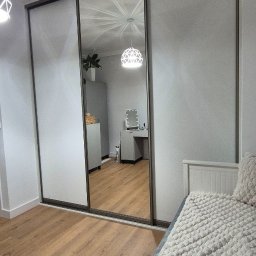 Verdini Andrzej Piotr - Trzydrzwiowa szafa wnękowa z lustrzanym frontem, odbijająca wnętrze pokoju z toaletką i szafką. Widoczny fragment łóżka z dekoracyjną narzutą.