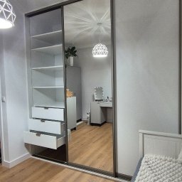 Verdini Andrzej Piotr - Szafa wnękowa z lustrzanymi drzwiami przesuwnymi, z otwartymi szufladami i półkami, odbijająca wnętrze pokoju z biurkiem i lampą. Widoczna część łóżka.