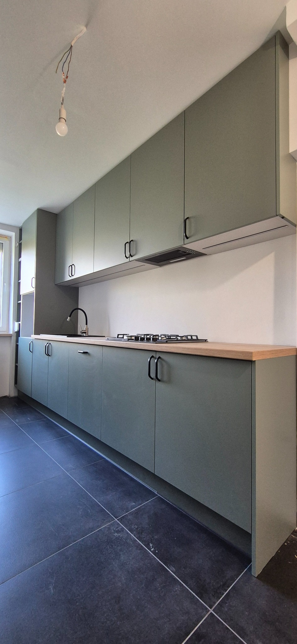 Nowoczesna kuchnia z oliwkowymi szafkami, drewnianym blatem i czarnymi uchwytami. Minimalistyczny design, czarne płytki podłogowe. Widoczne niedokończone oświetlenie.