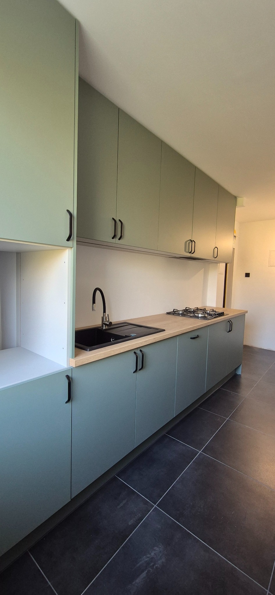 Nowoczesna kuchnia z szafkami w odcieniach zieleni, czarnym zlewem i drewnianym blatem. Minimalistyczne uchwyty i ciemna podłoga dodają elegancji.
