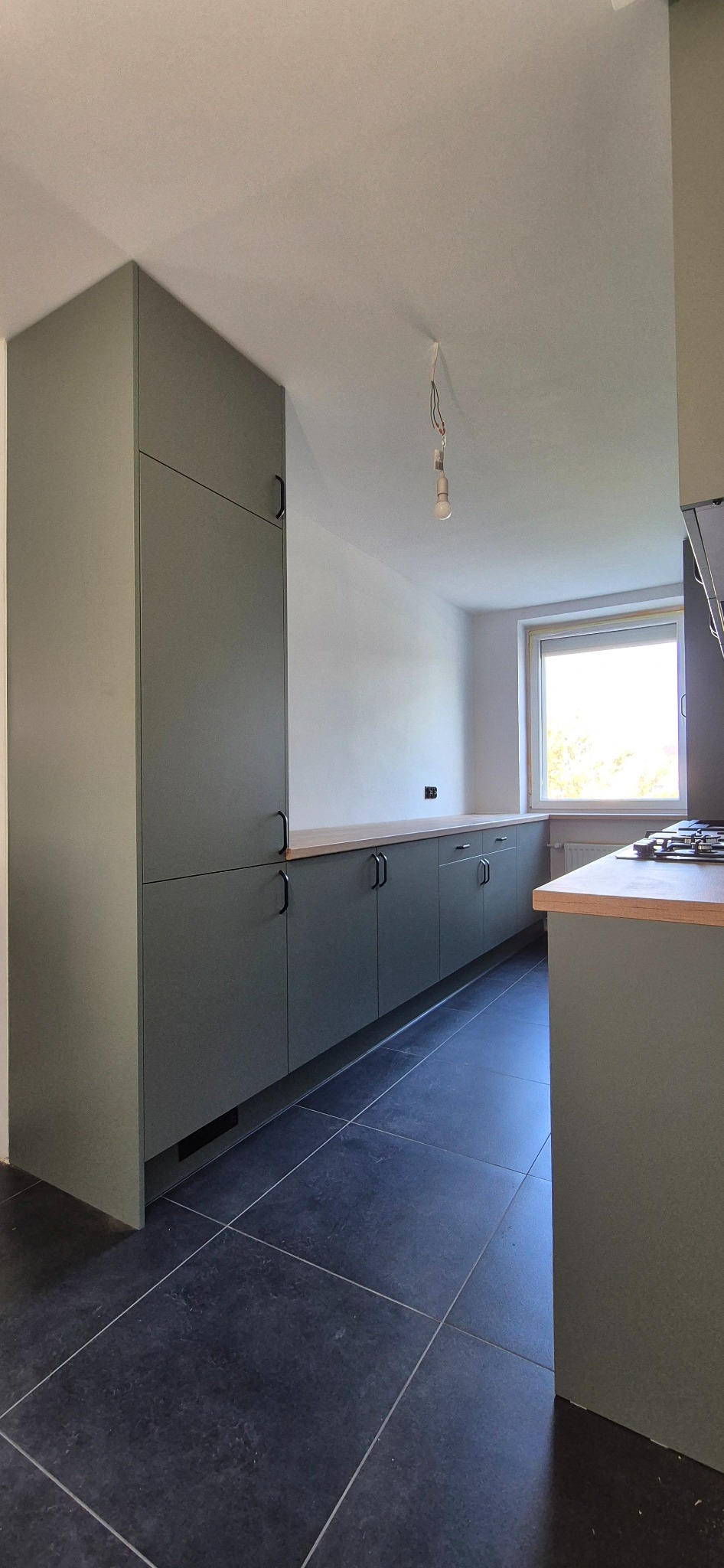 Nowoczesna kuchnia z oliwkowymi szafkami i drewnianym blatem, czarne płytki podłogowe, minimalistyczny design, surowa żarówka zwisająca z sufitu.