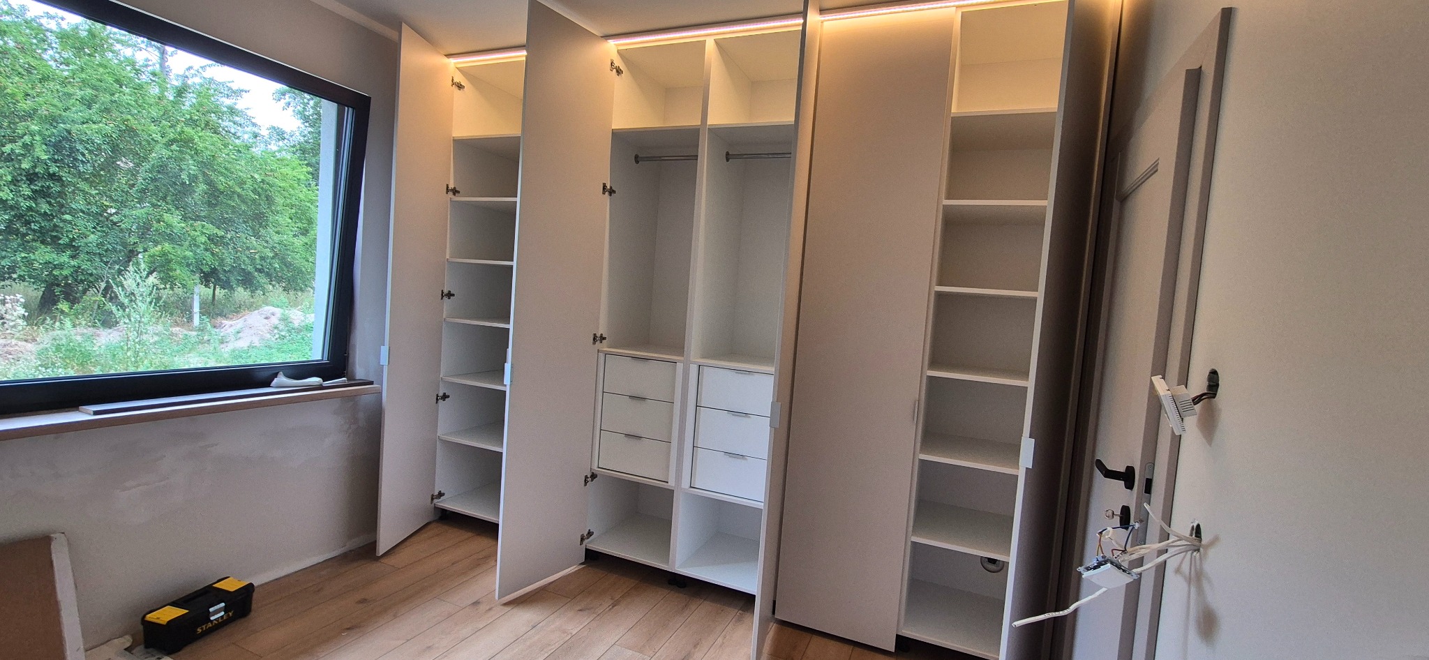 Jasna szafa wnękowa z oświetleniem LED, otwartymi półkami i szufladami, widoczna z boku okno z zielenią. Na podłodze skrzynka narzędziowa.