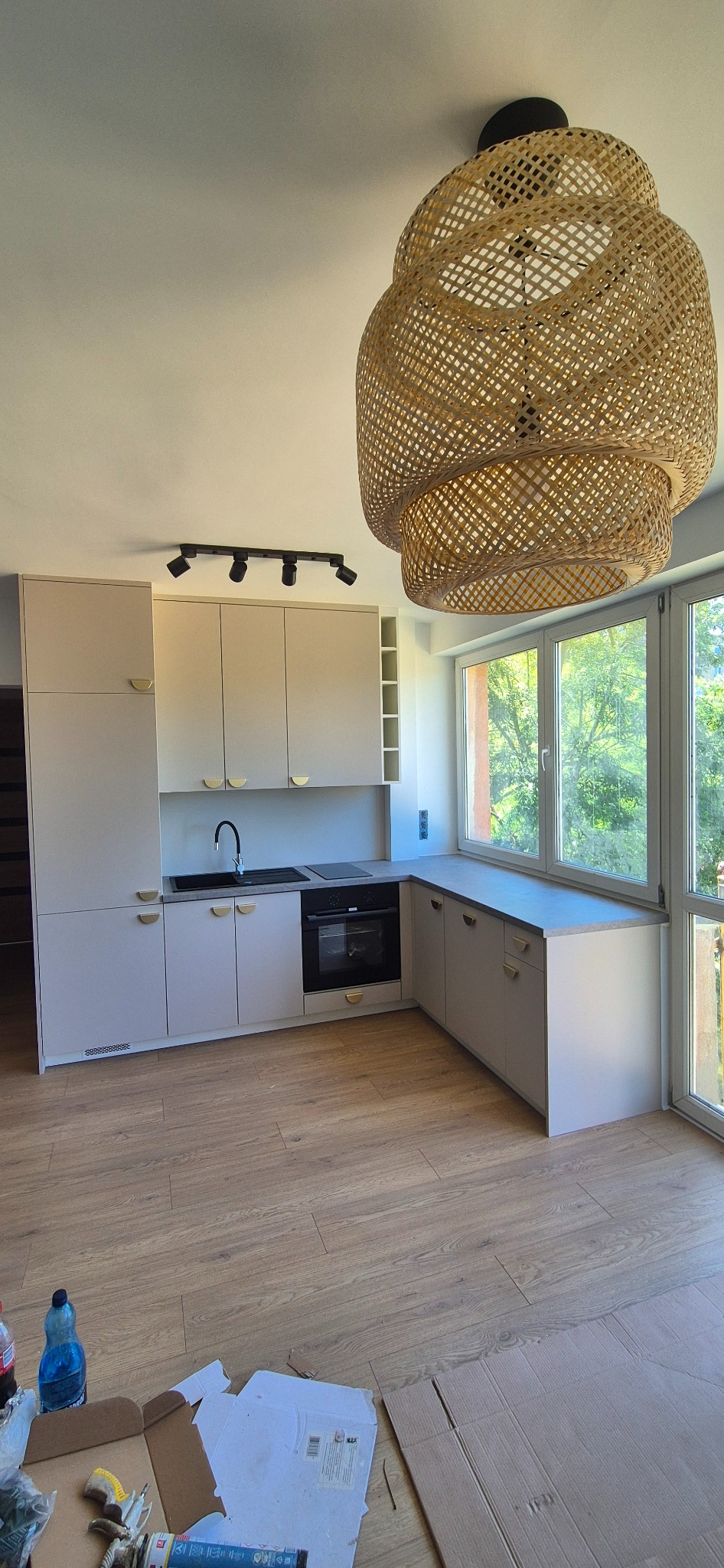 Jasna kuchnia z beżowymi frontami, złotymi uchwytami i dużym oknem. W tle widoczny ażurowy, okrągły abażur. Na podłodze kartony i butelka.