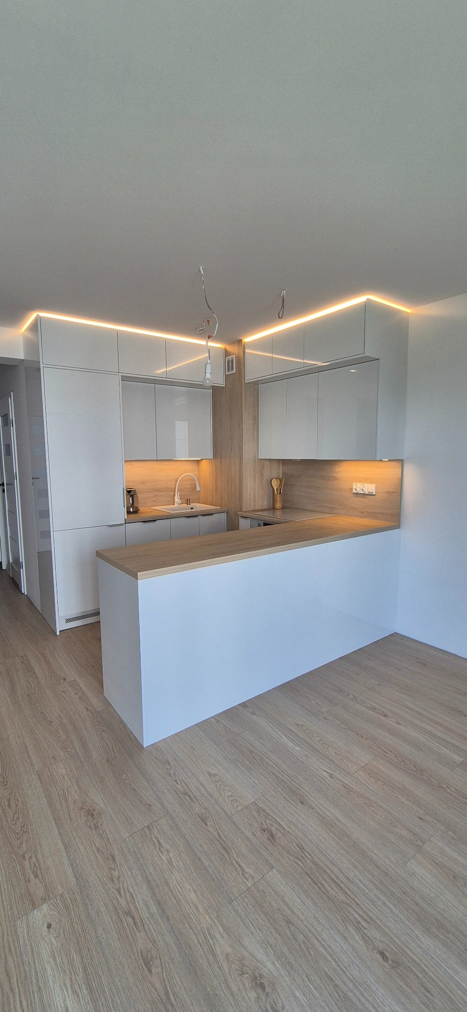 Nowoczesna, jasna kuchnia z białymi szafkami i drewnianym blatem, oświetlona listwami LED. Widoczne niedokończone podłączenie lampy sufitowej.
