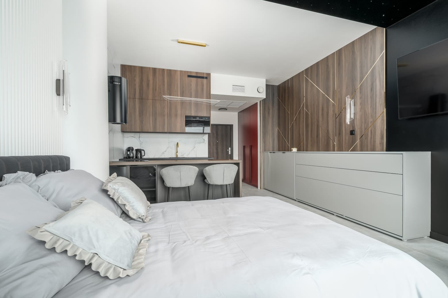Nowoczesne studio: łóżko z szarymi poduszkami, aneks kuchenny z drewnianymi szafkami i marmurowym blatem, szare krzesła barowe, ściana z drewnianą okładziną i złotymi akcentami.
