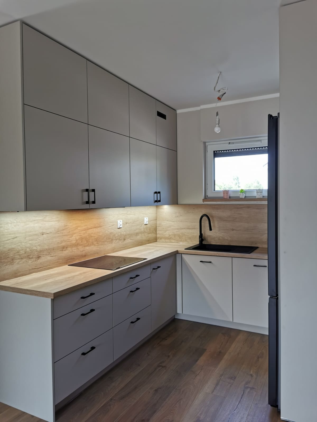 Nowoczesna kuchnia z szarymi szafkami, drewnianym blatem i czarnymi uchwytami. Widoczna płyta indukcyjna i czarna bateria. Podświetlenie LED pod szafkami górnymi.