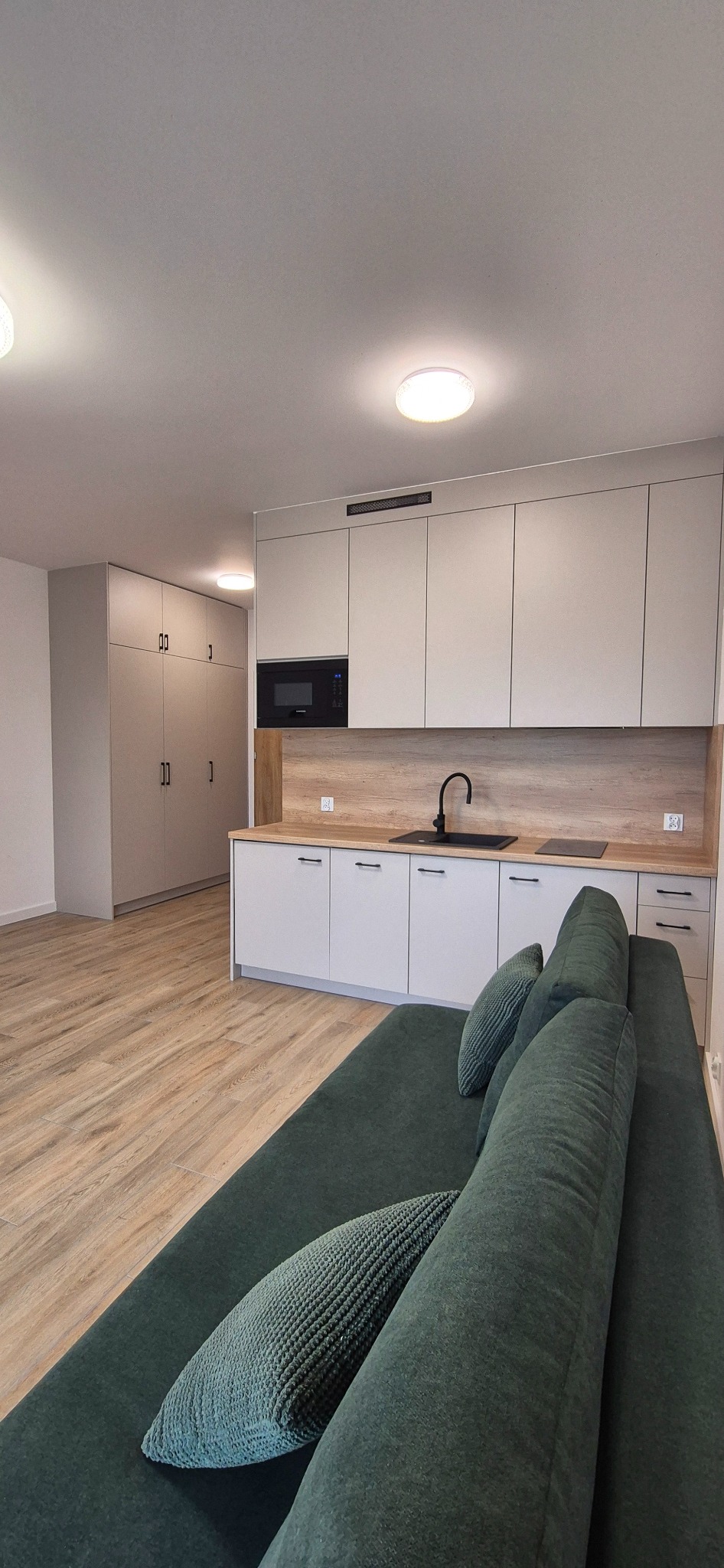 Jasna kuchnia z drewnianym blatem i czarną armaturą, szafa wnękowa w tle, widok z perspektywy sofy z zielonymi poduszkami. Nowoczesne wykończenie.