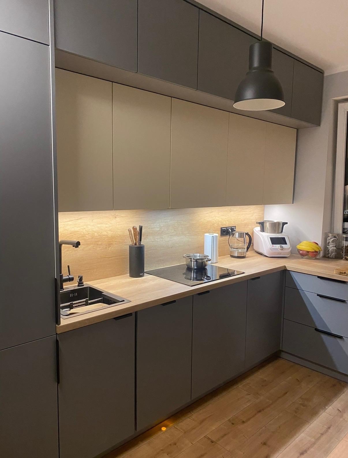 Nowoczesna kuchnia z szarymi szafkami, drewnianym blatem i oświetleniem LED pod szafkami wiszącymi. Na blacie czajnik, robot kuchenny i akcesoria.